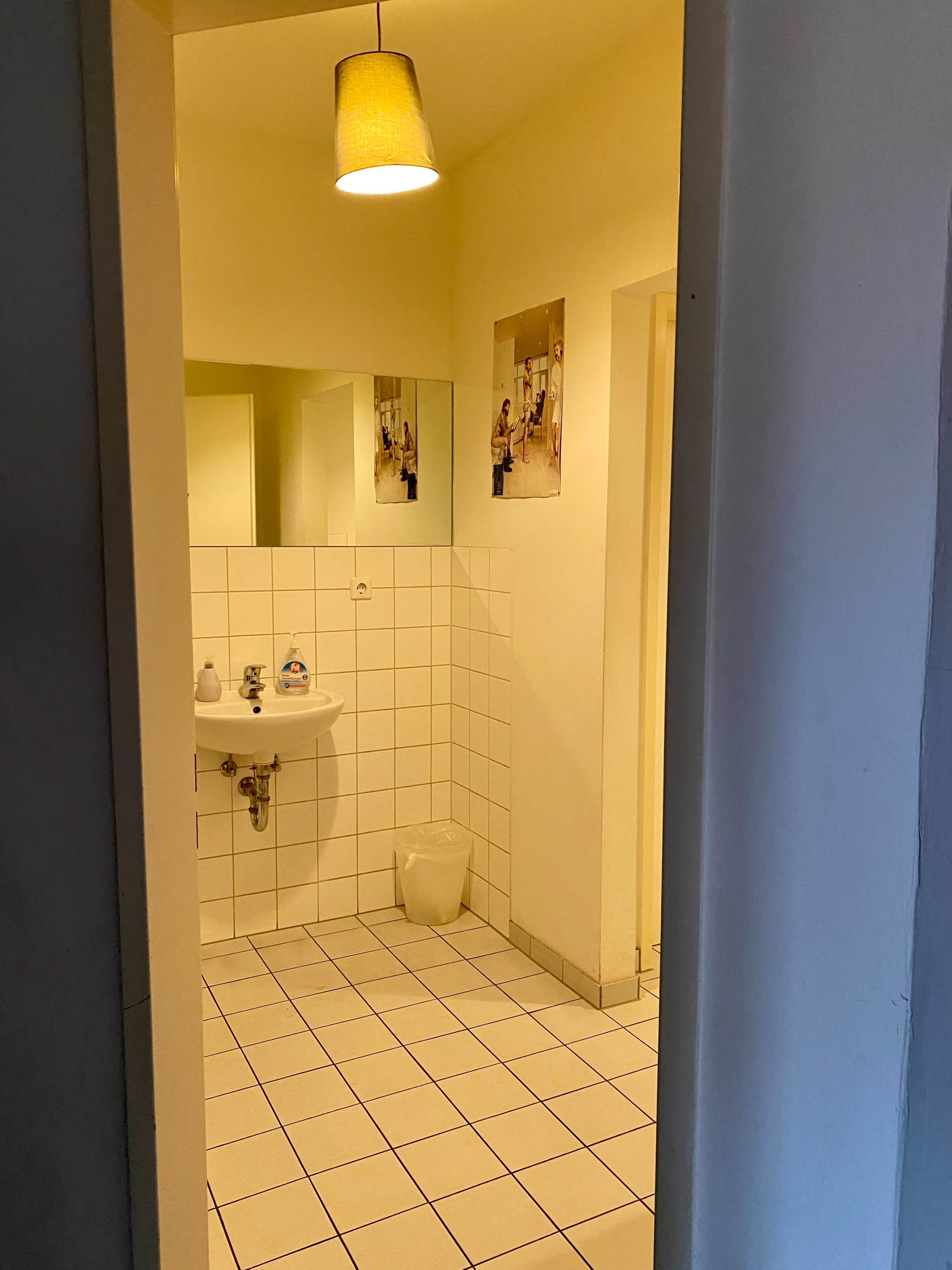 A small bathroom with white tiled walls, a sink, a light fixture, and a wastebasket in the corner.