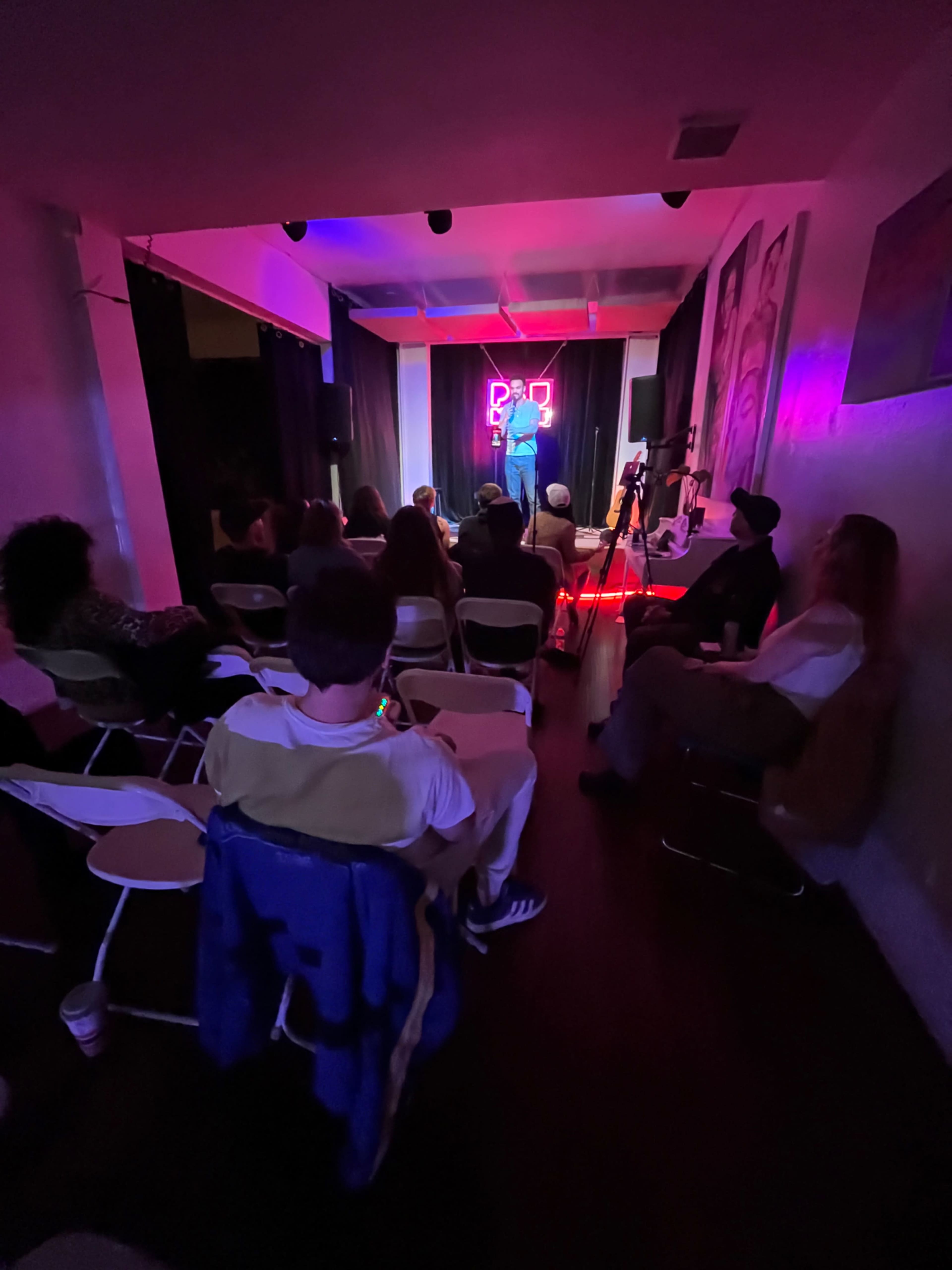 A performer stands on stage with neon lights in a small venue while an audience watches from seated rows.