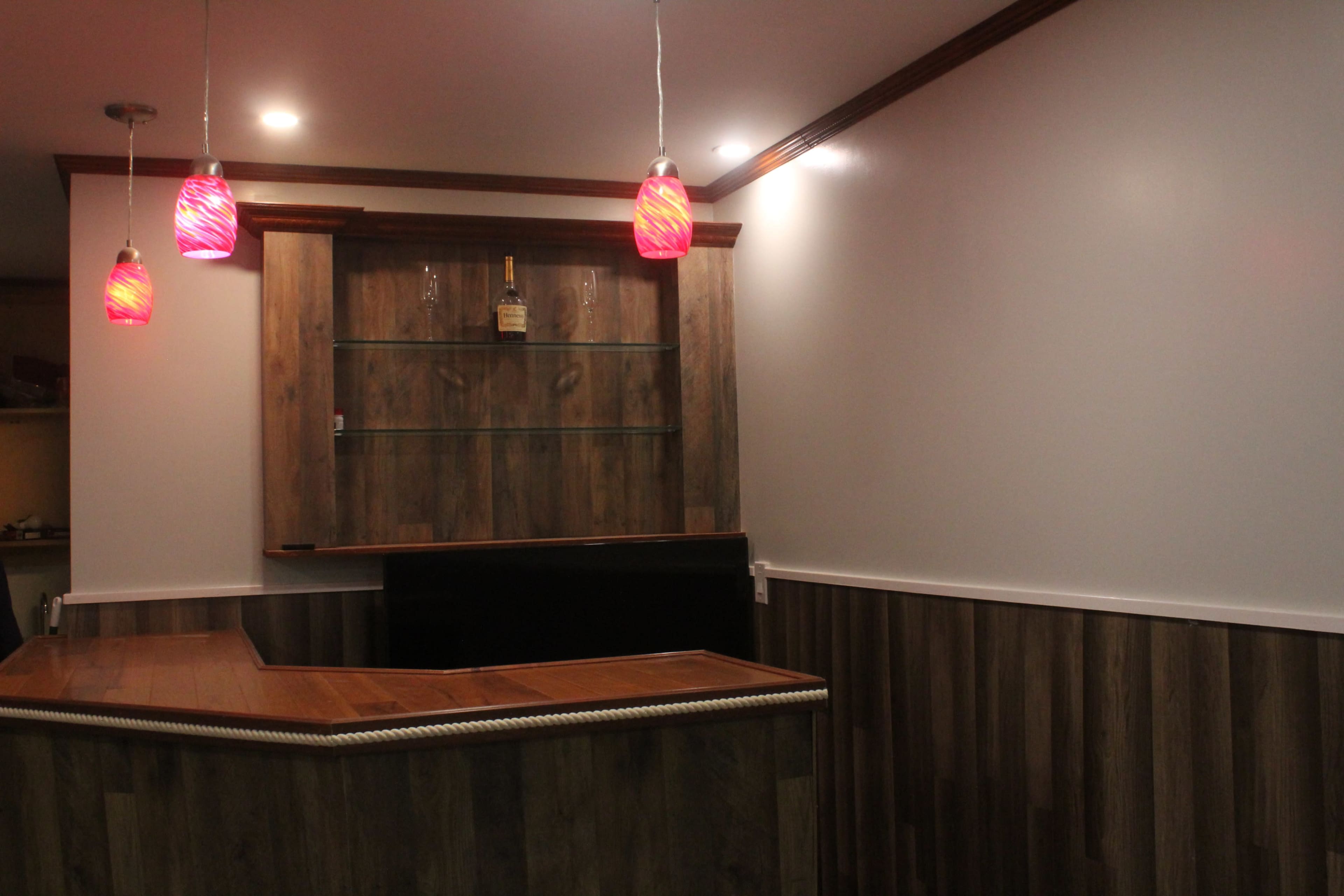 The image shows a bar area featuring a wooden countertop, glass shelves, and pendant lights with pink shades.