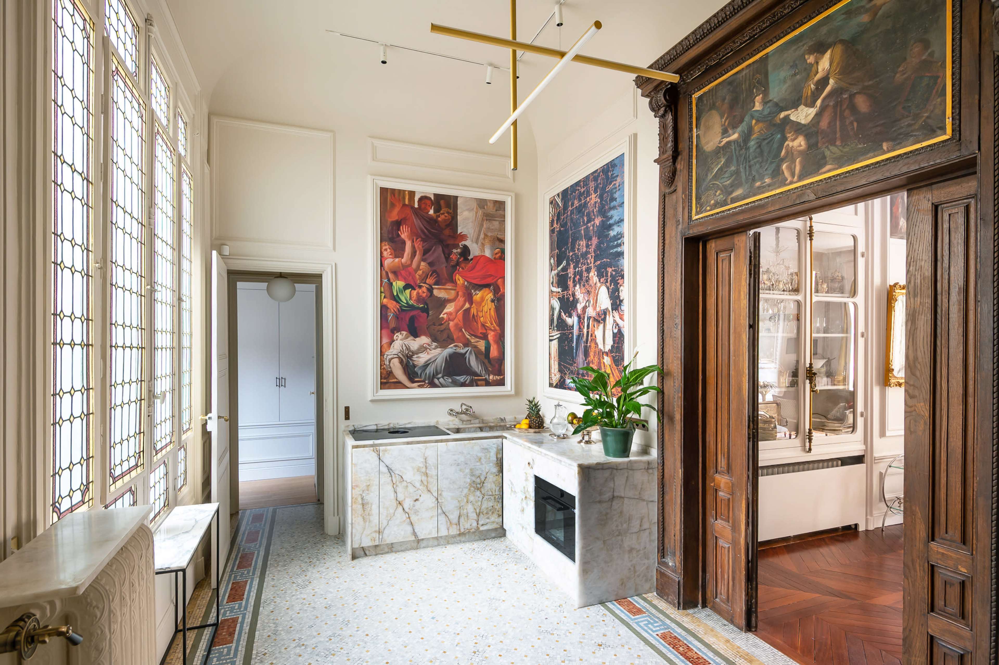 The image depicts a bright kitchen featuring marble countertops and a large window with stained glass, adjacent to an ornate wooden doorway leading to a dining area.