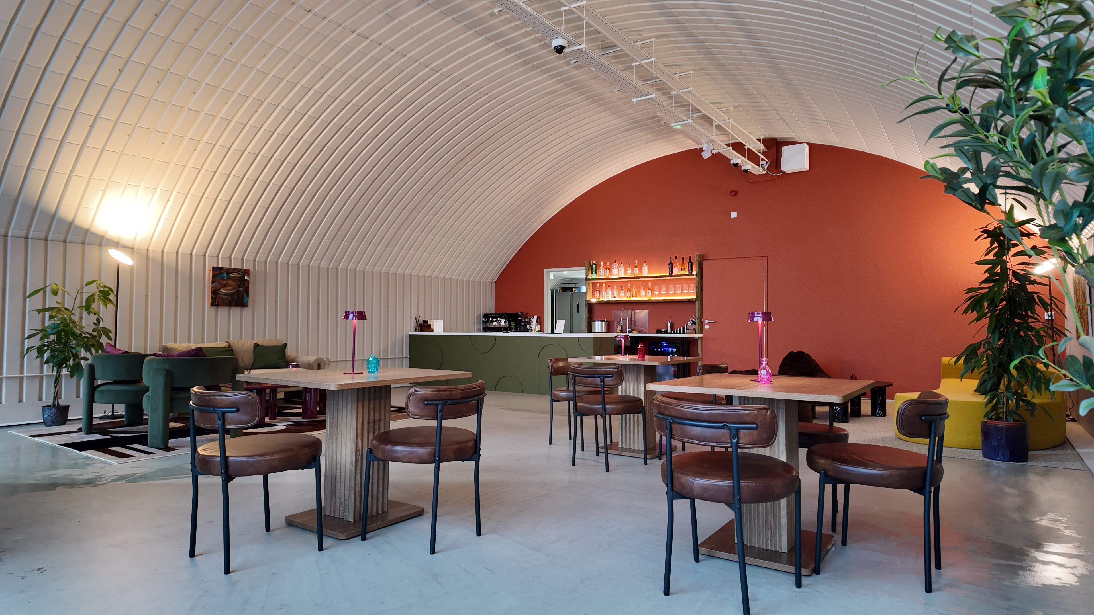 The image shows a modern, minimalist cafe with an arched ceiling, wooden tables, and seating arranged around a bar area with soft lighting.