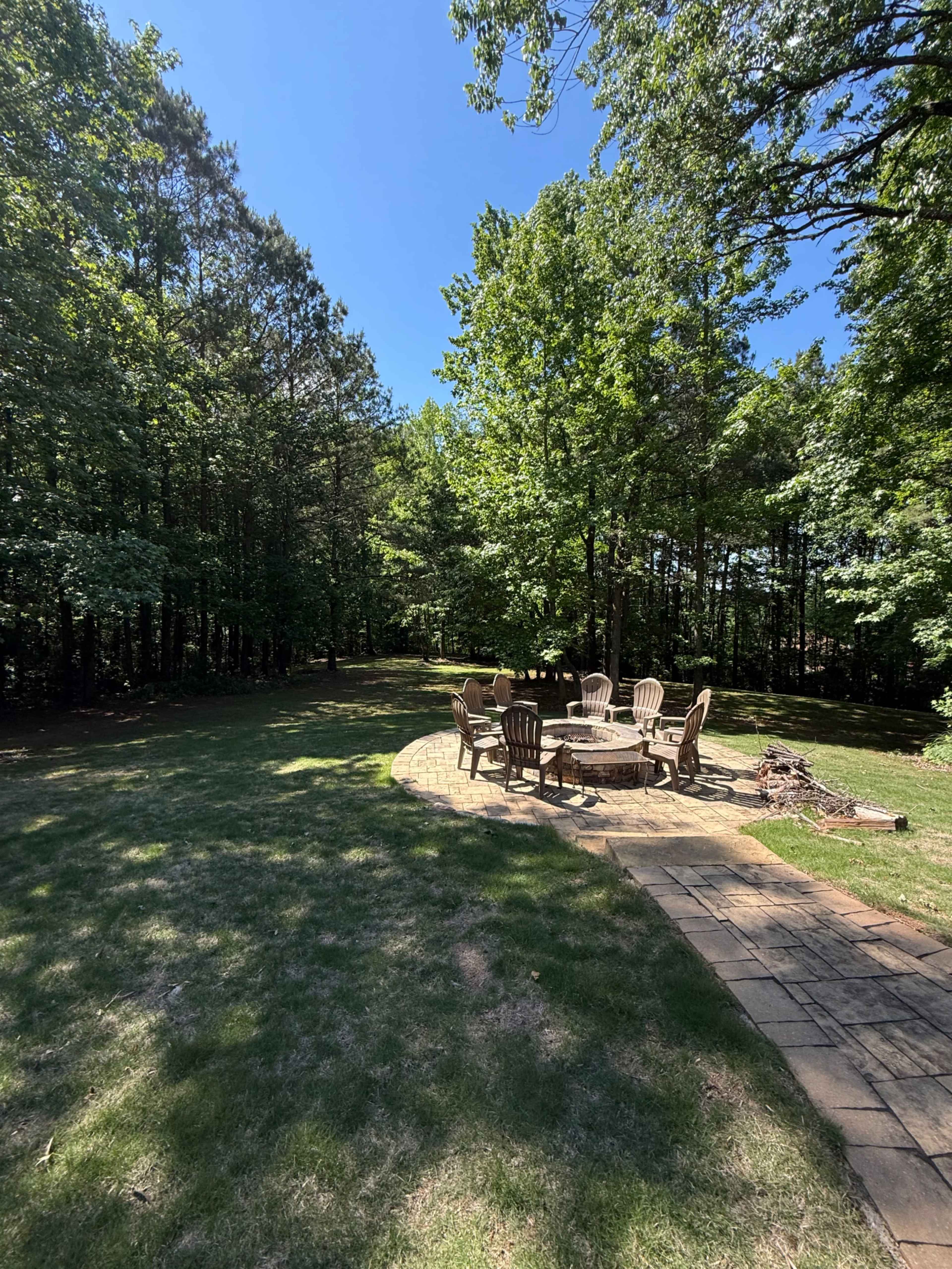 A stone patio with a circular arrangement of wooden chairs sits in a clearing surrounded by trees.