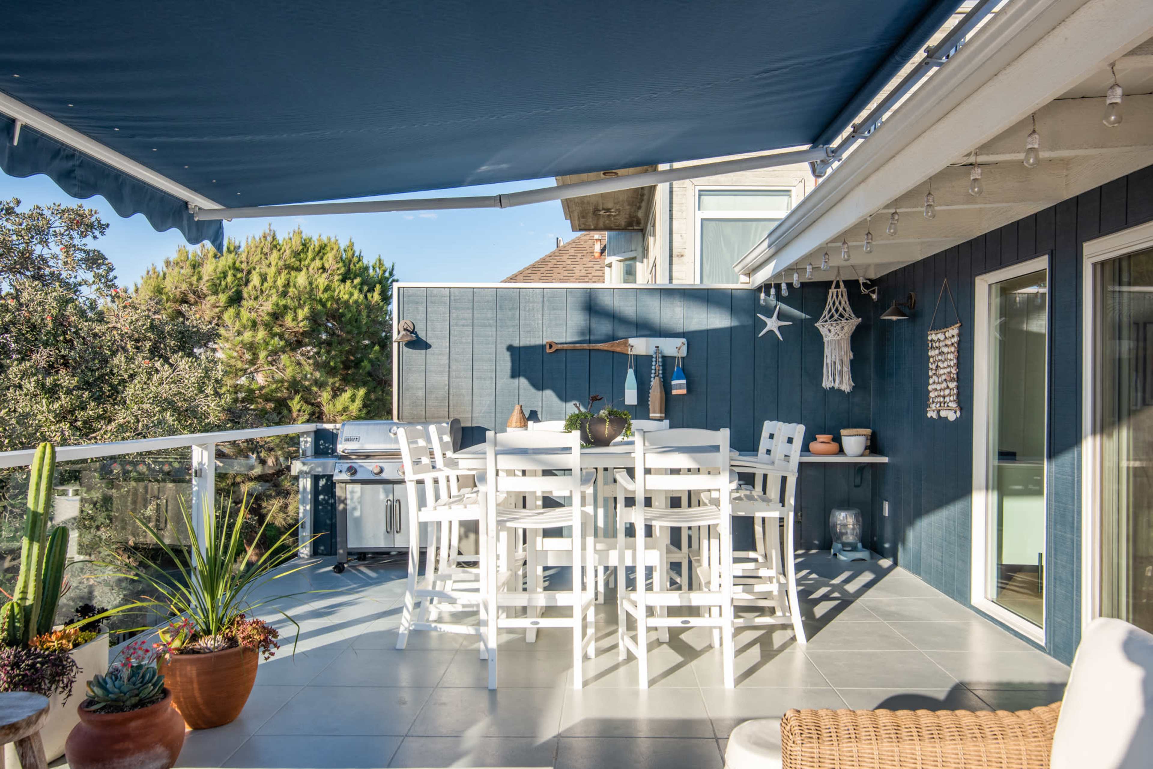 A patio area with a white outdoor dining set, a barbecue grill, and decorative plants under a blue canopy.