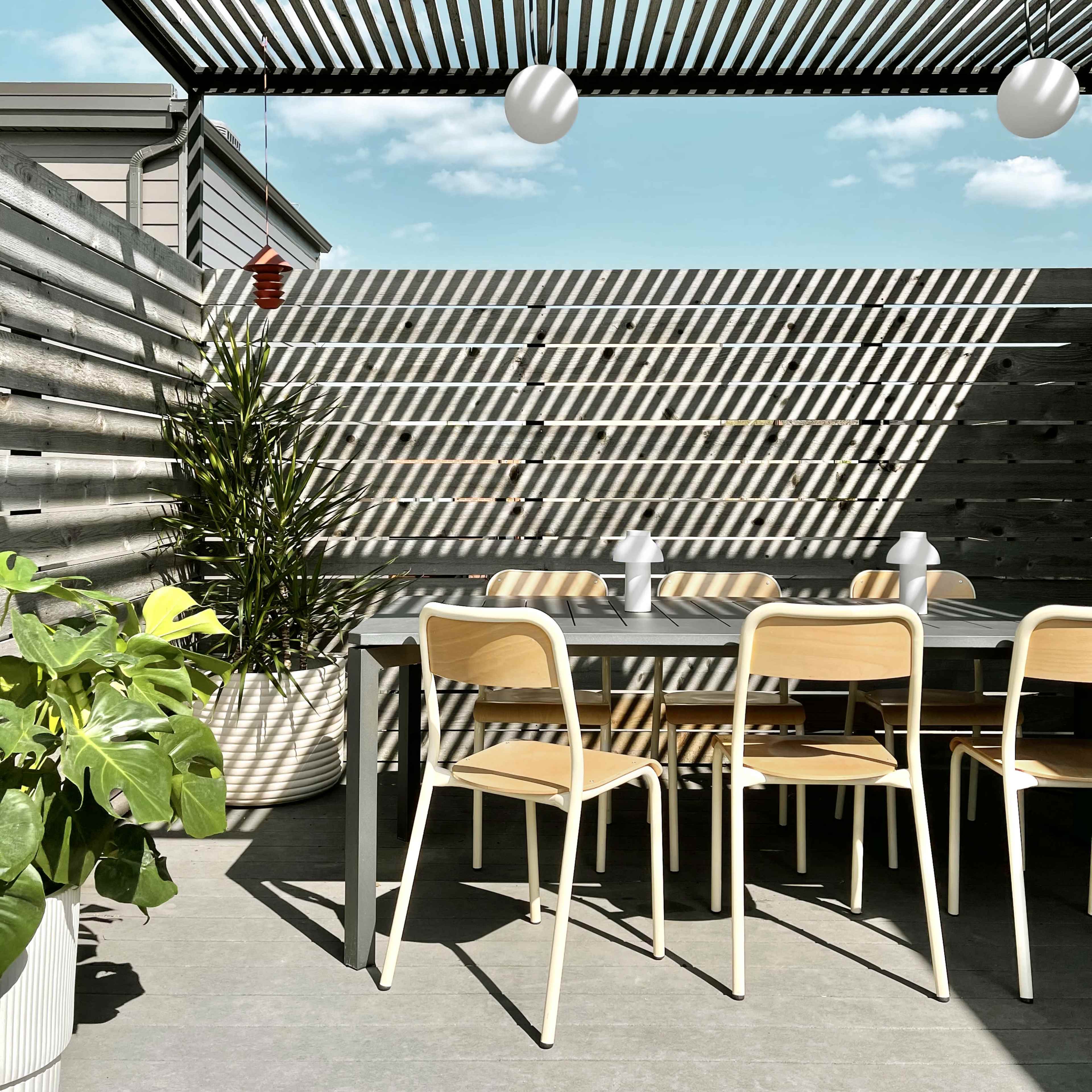 A modern outdoor dining area features a table with several wooden chairs, surrounded by bamboo plants and shaded by a slatted pergola.