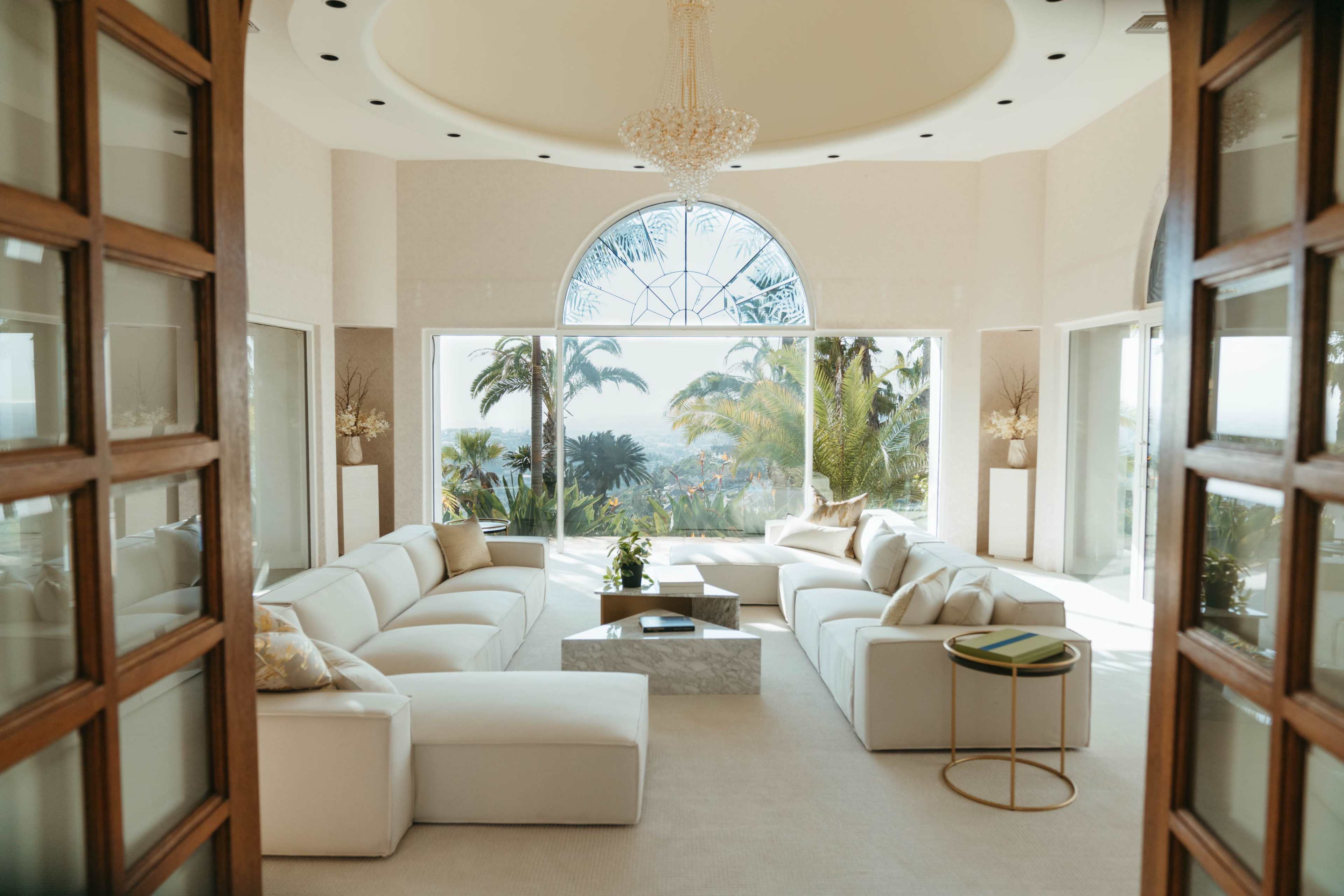 A spacious, bright living room features a large curved window with a chandelier overhead and modern white seating arranged around a coffee table.