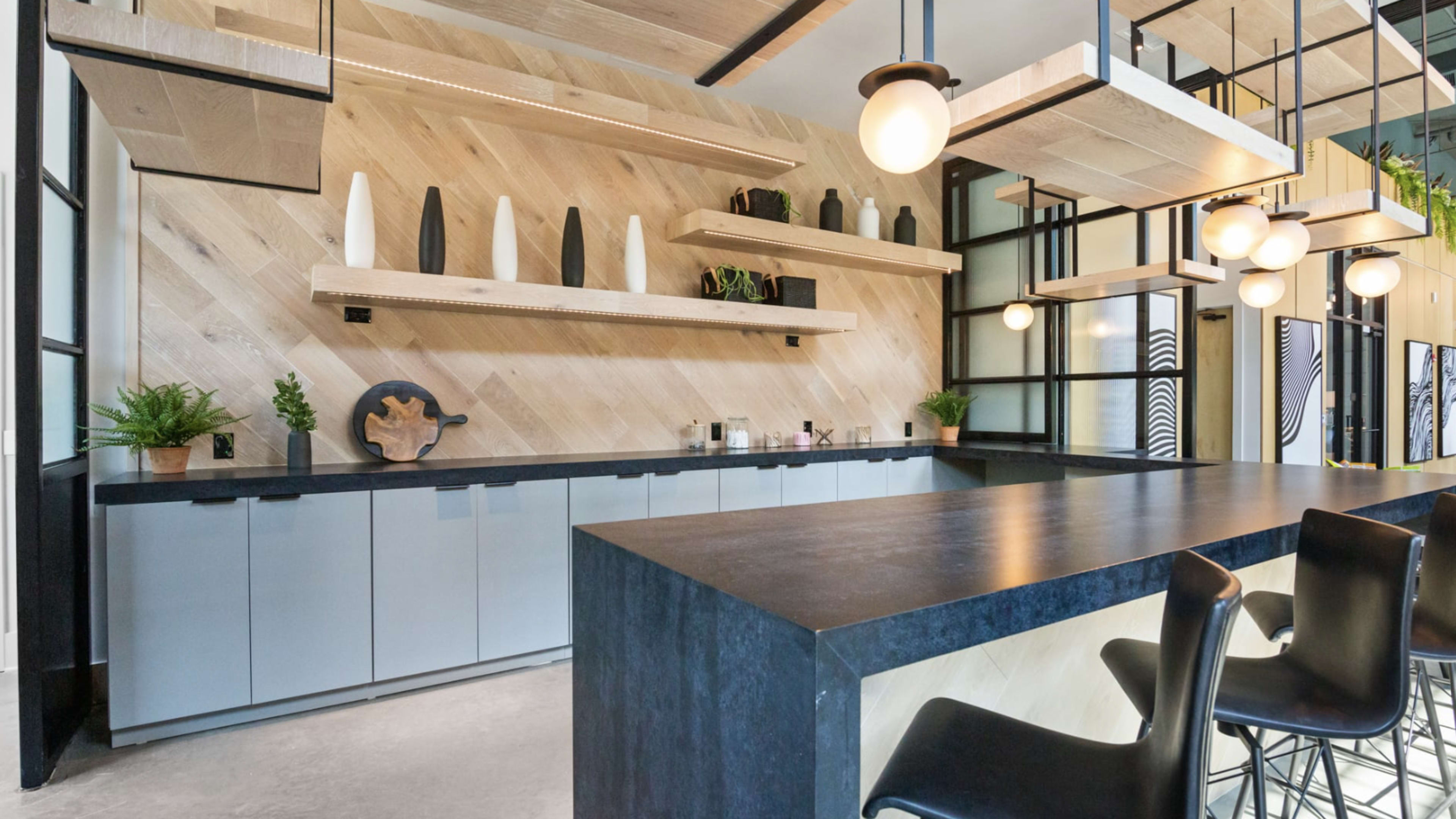 The image shows a modern kitchen with a large black countertop, wooden cabinetry, and decorative shelves featuring plants and vases.