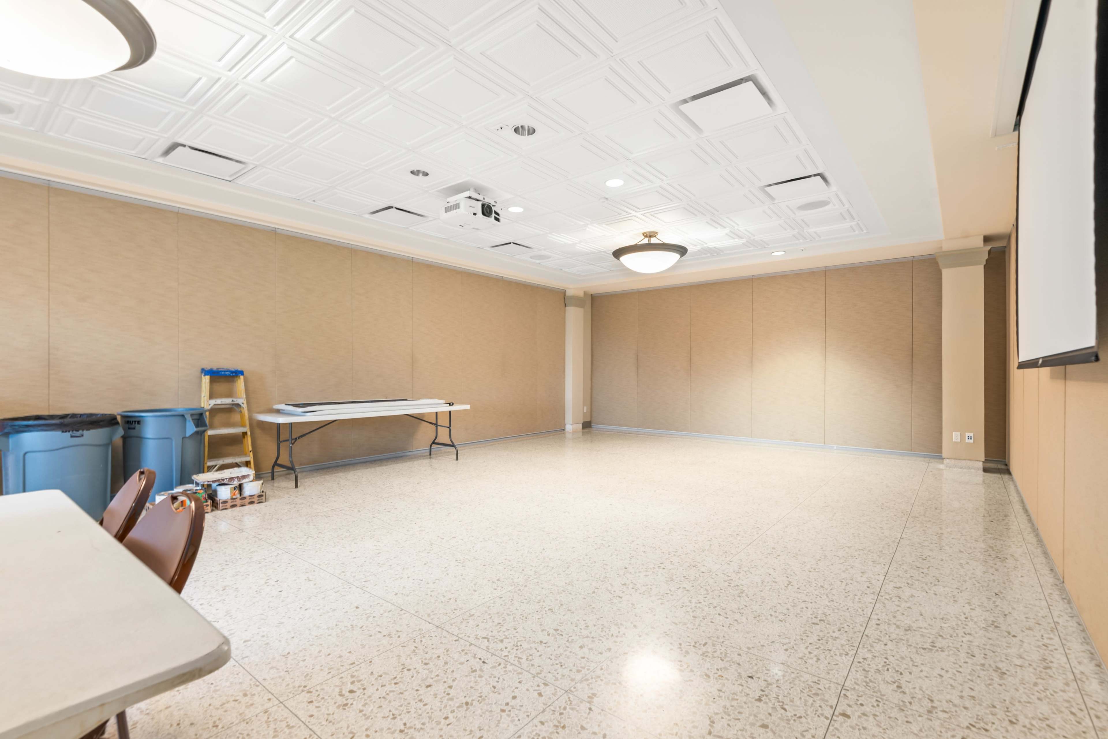 The image shows a large, empty meeting room with beige walls, a projector on the ceiling, and a table and chairs positioned against one side.
