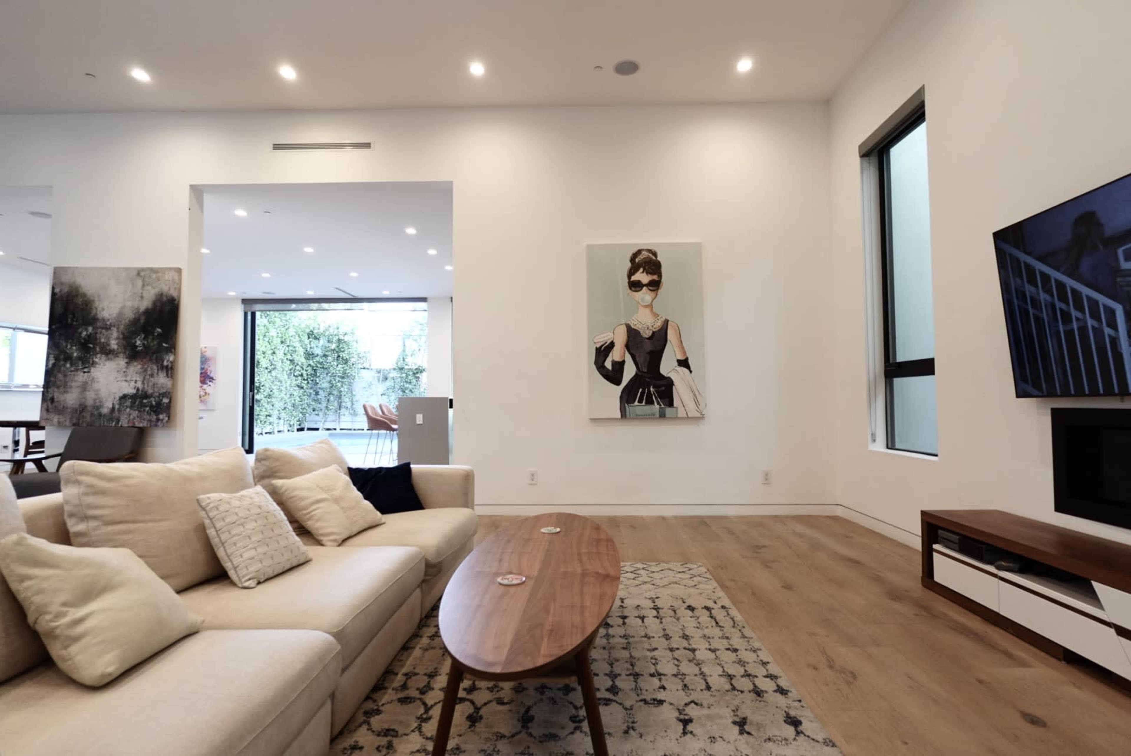 The living room features a light-colored sofa, a wooden coffee table, and a large artwork on the wall, with large windows showing a glimpse of an outdoor space.