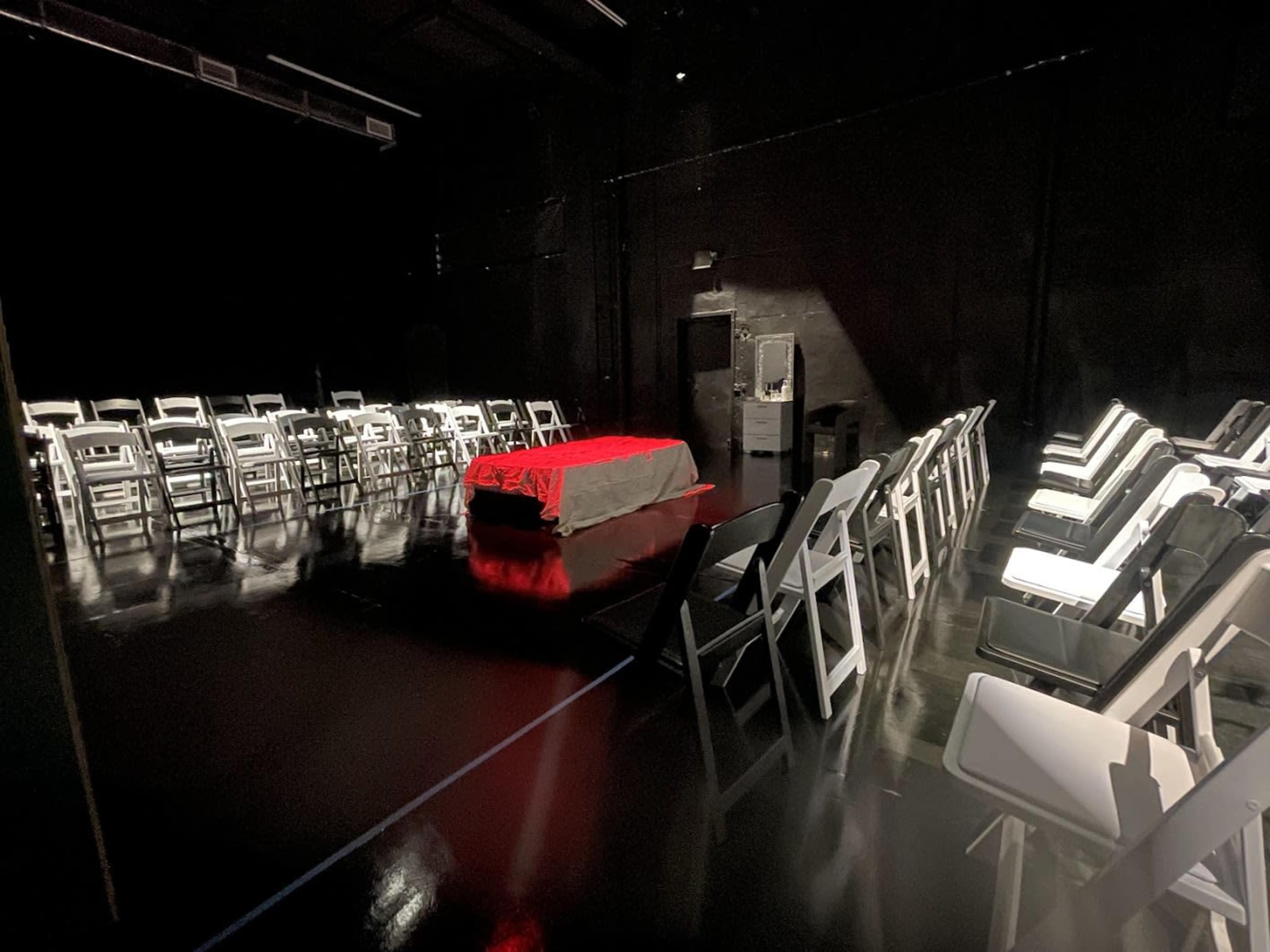 A dark room features a table covered with a red cloth at the center, surrounded by white folding chairs arranged in two semicircles.
