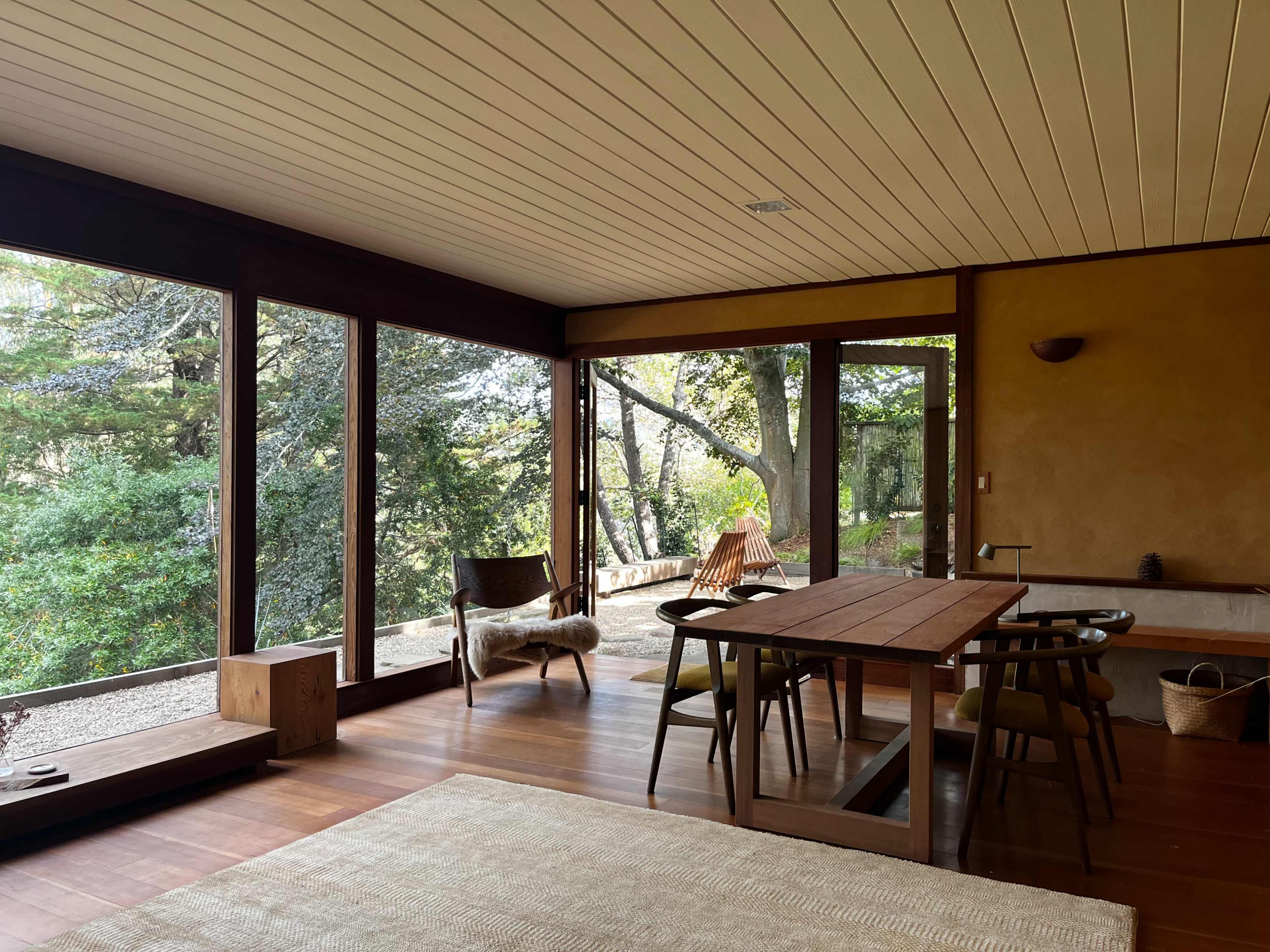 A modern interior features large windows overlooking a wooded area, with a wooden dining table and simple furnishings.