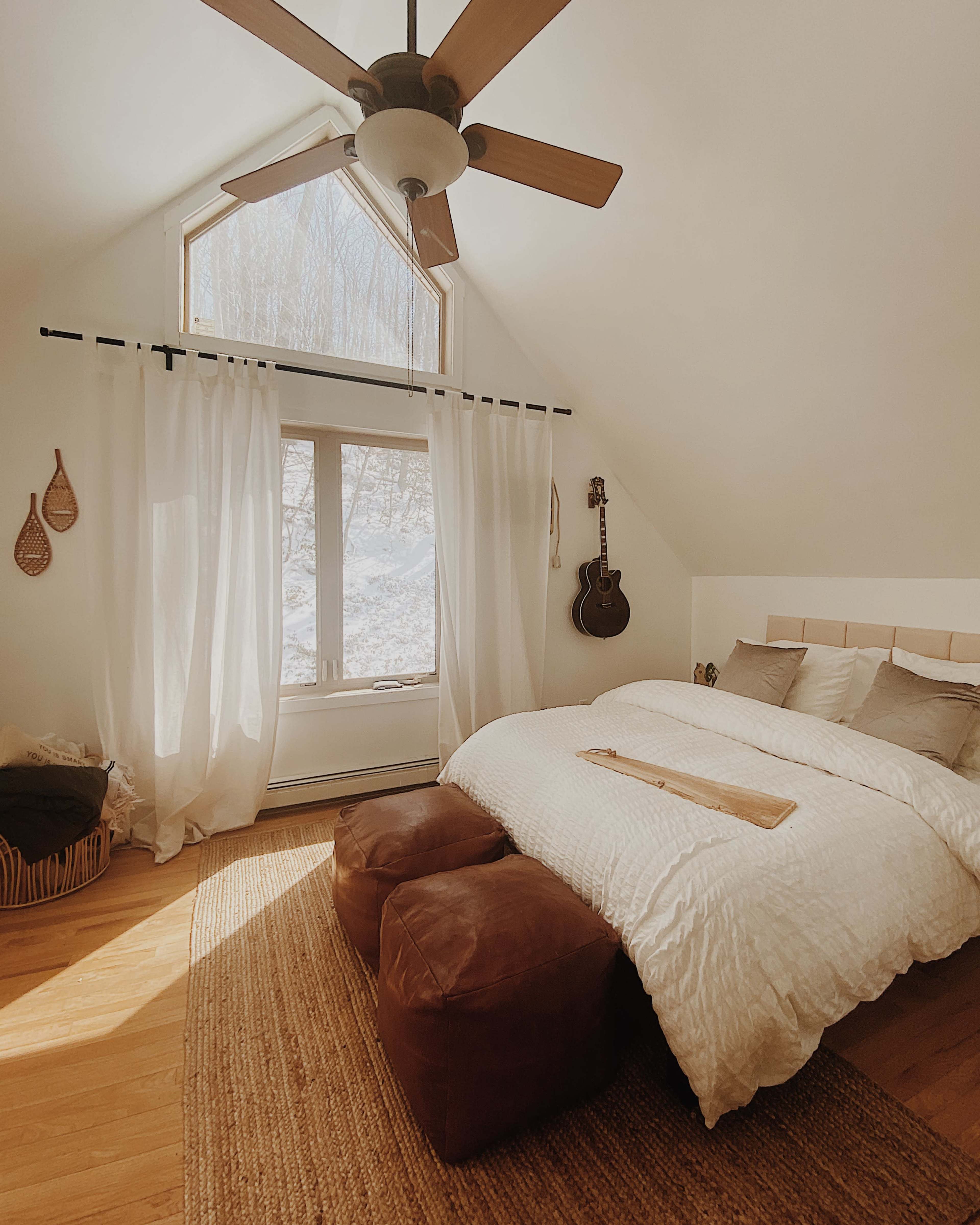 A cozy bedroom features a large window with white curtains, a bed with a neutral-colored bedspread, and a ceiling fan, along with a guitar hanging on the wall.