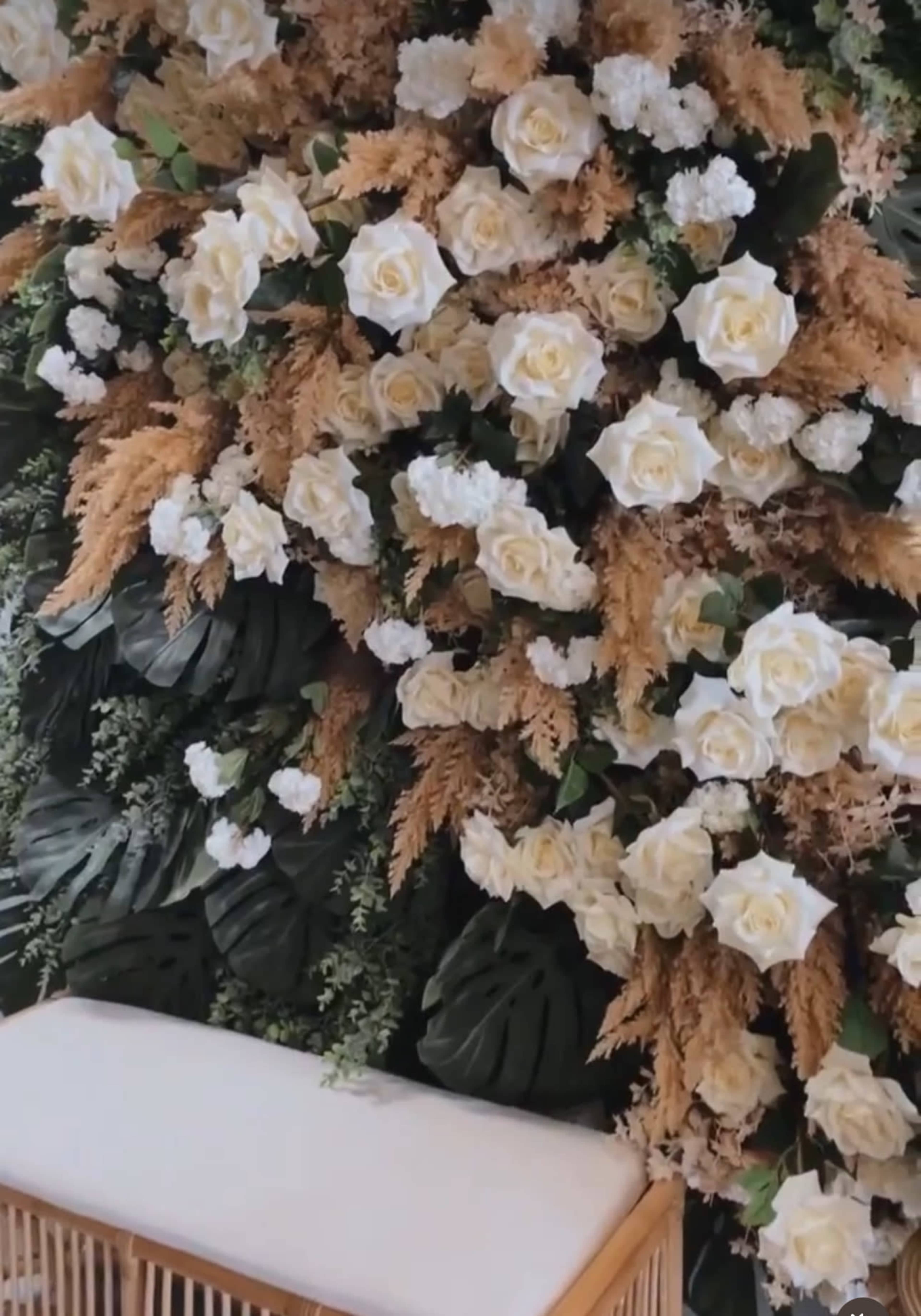 The image shows a wall decorated with white roses and green foliage, alongside dried plants, with a wooden bench at the bottom.