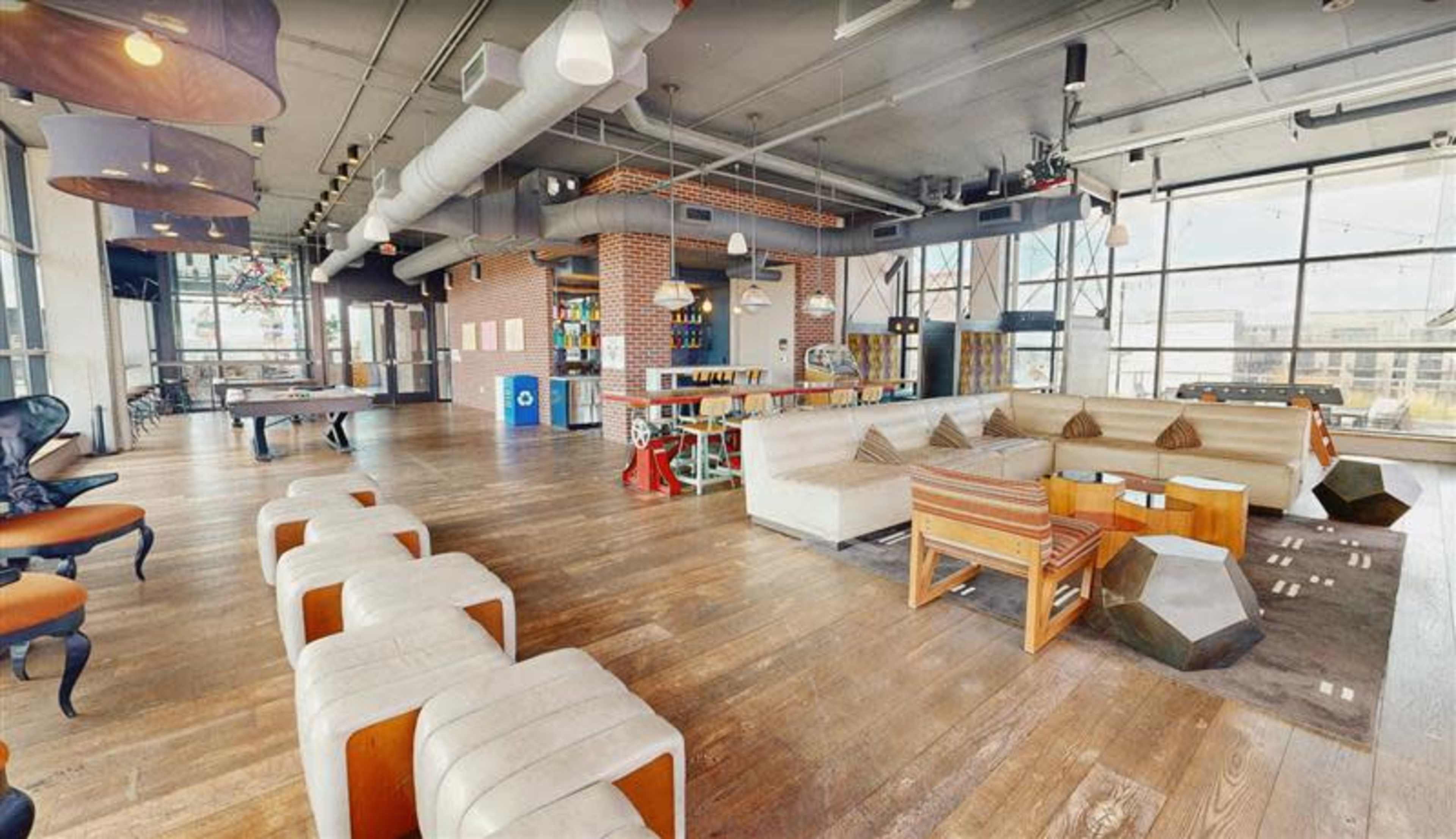 The image shows a spacious, modern lounge area with various seating arrangements, including couches and stools, alongside a game table and shelves filled with items.