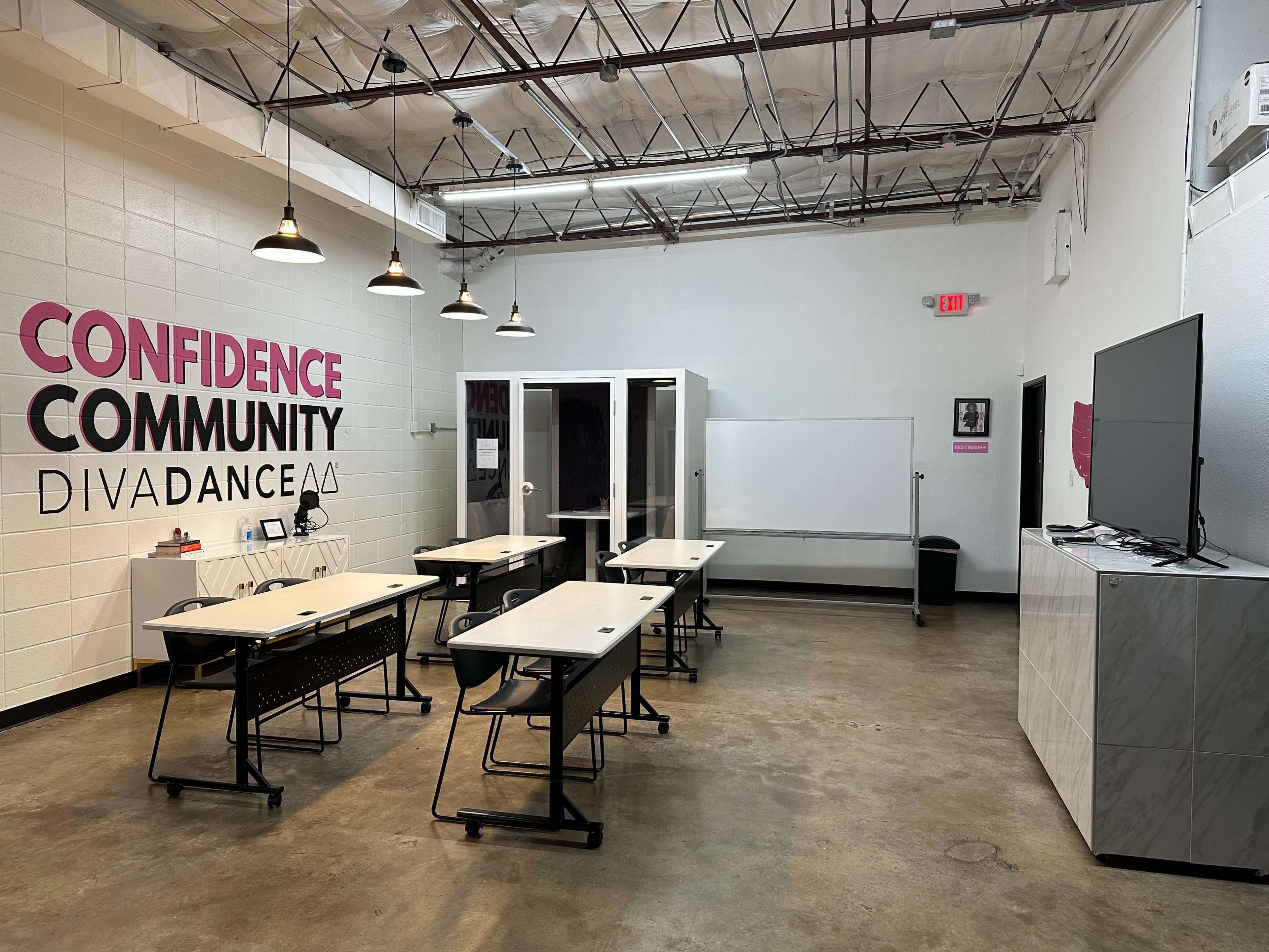 A spacious classroom with several desks arranged in rows, a whiteboard, and a large TV on the wall, with the words "Confidence, Community, DivaDance" displayed prominently.