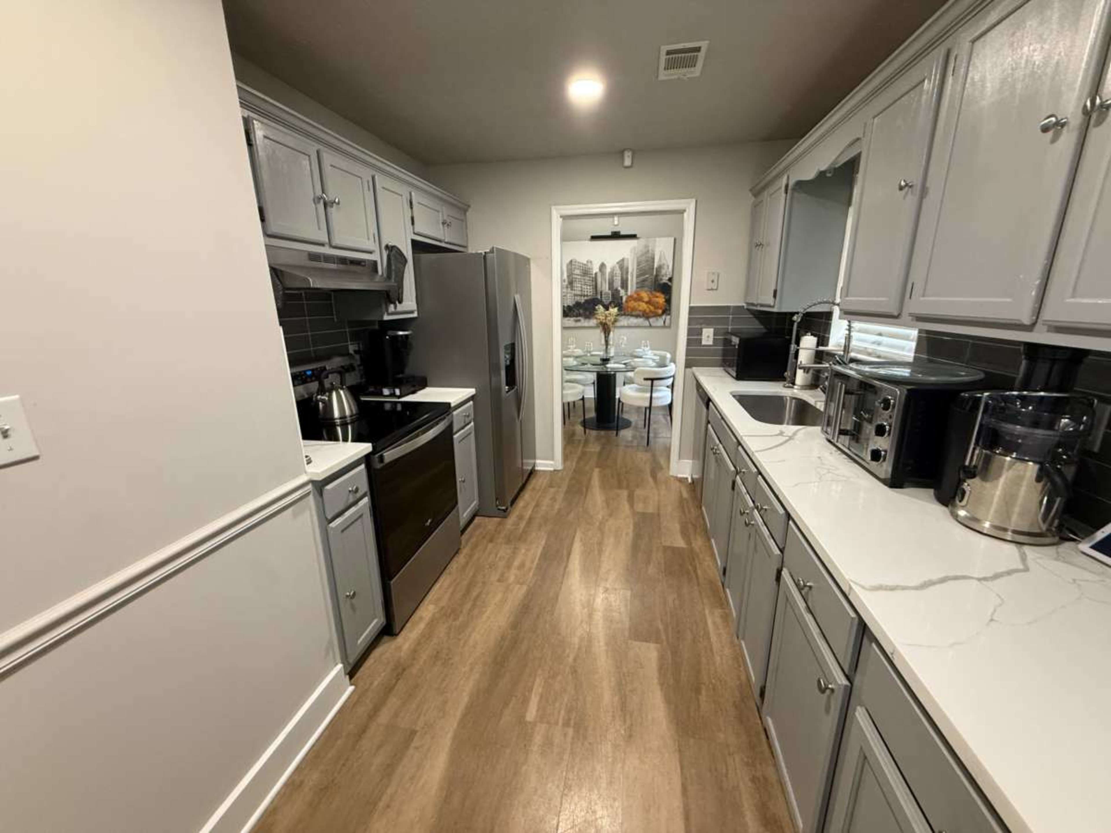 The image shows a modern kitchen with gray cabinetry, stainless steel appliances, and a doorway leading to a dining area with a table and chairs.