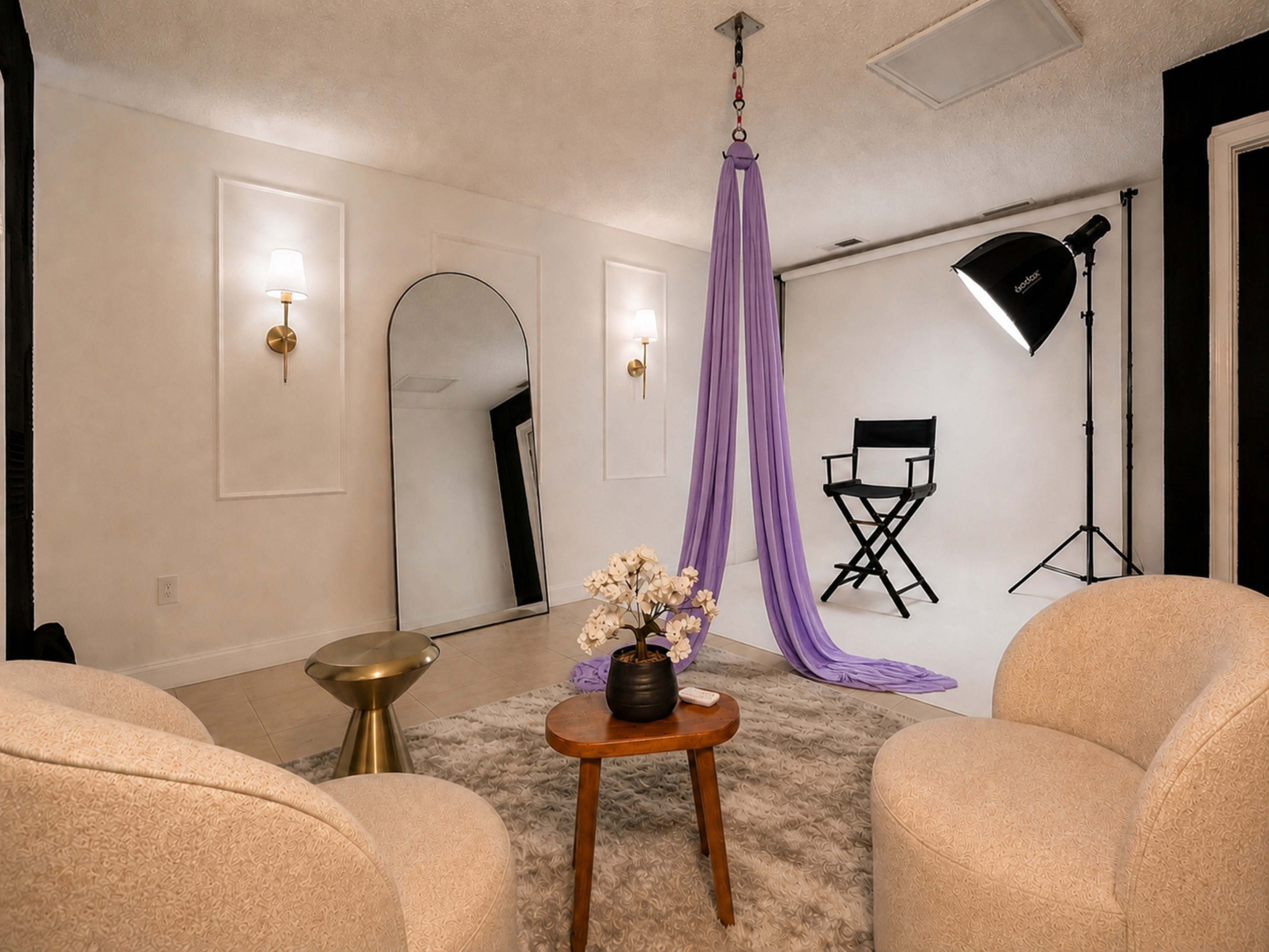 The image features a well-lit studio space with two upholstered chairs, a small wooden table, a large mirror, a hanging fabric drape, and a photography setup including a light and a director's chair.