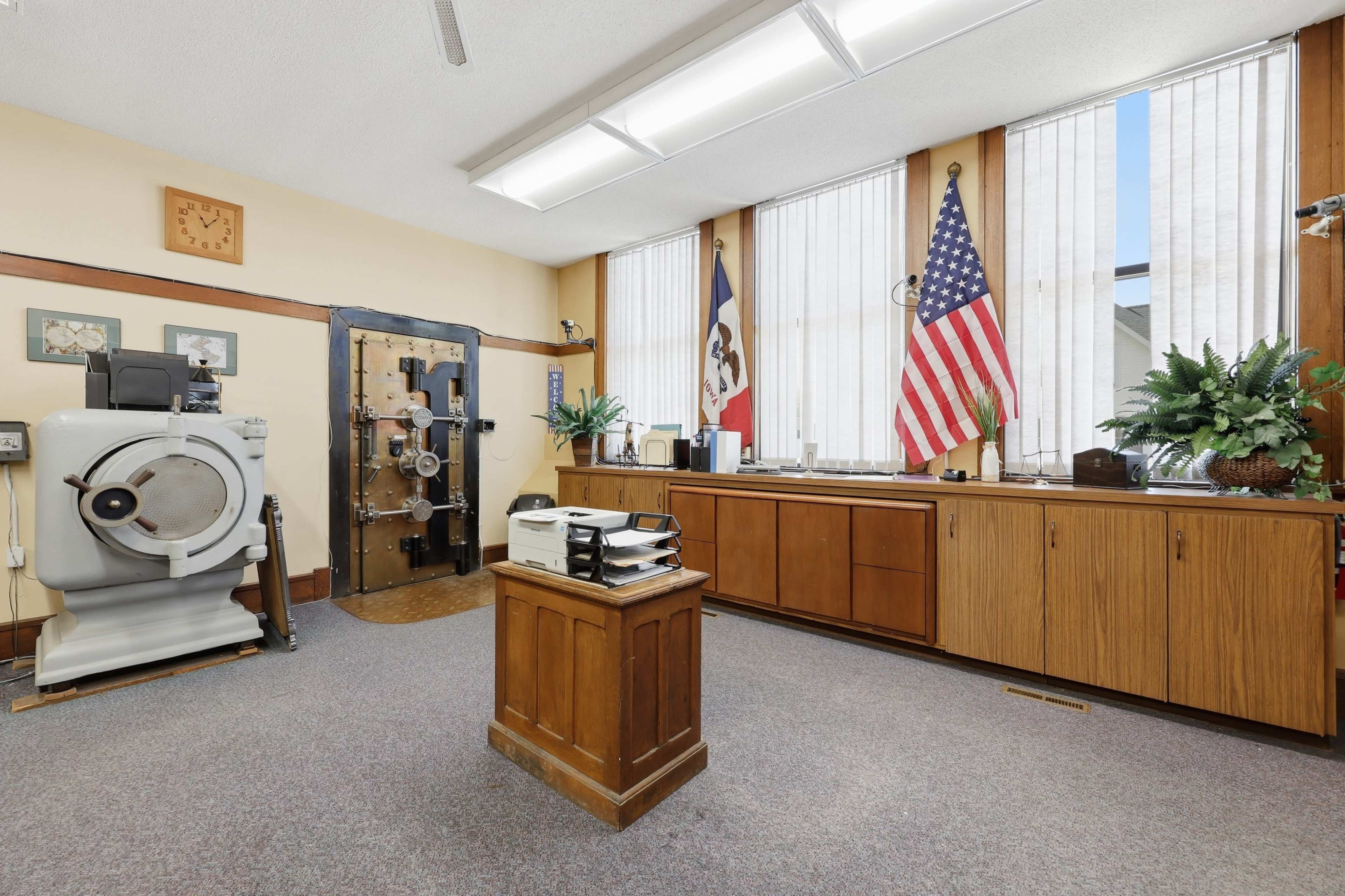 The image shows an interior room featuring a large safe, two windows with blinds, American and Iowa state flags, and wooden cabinets.