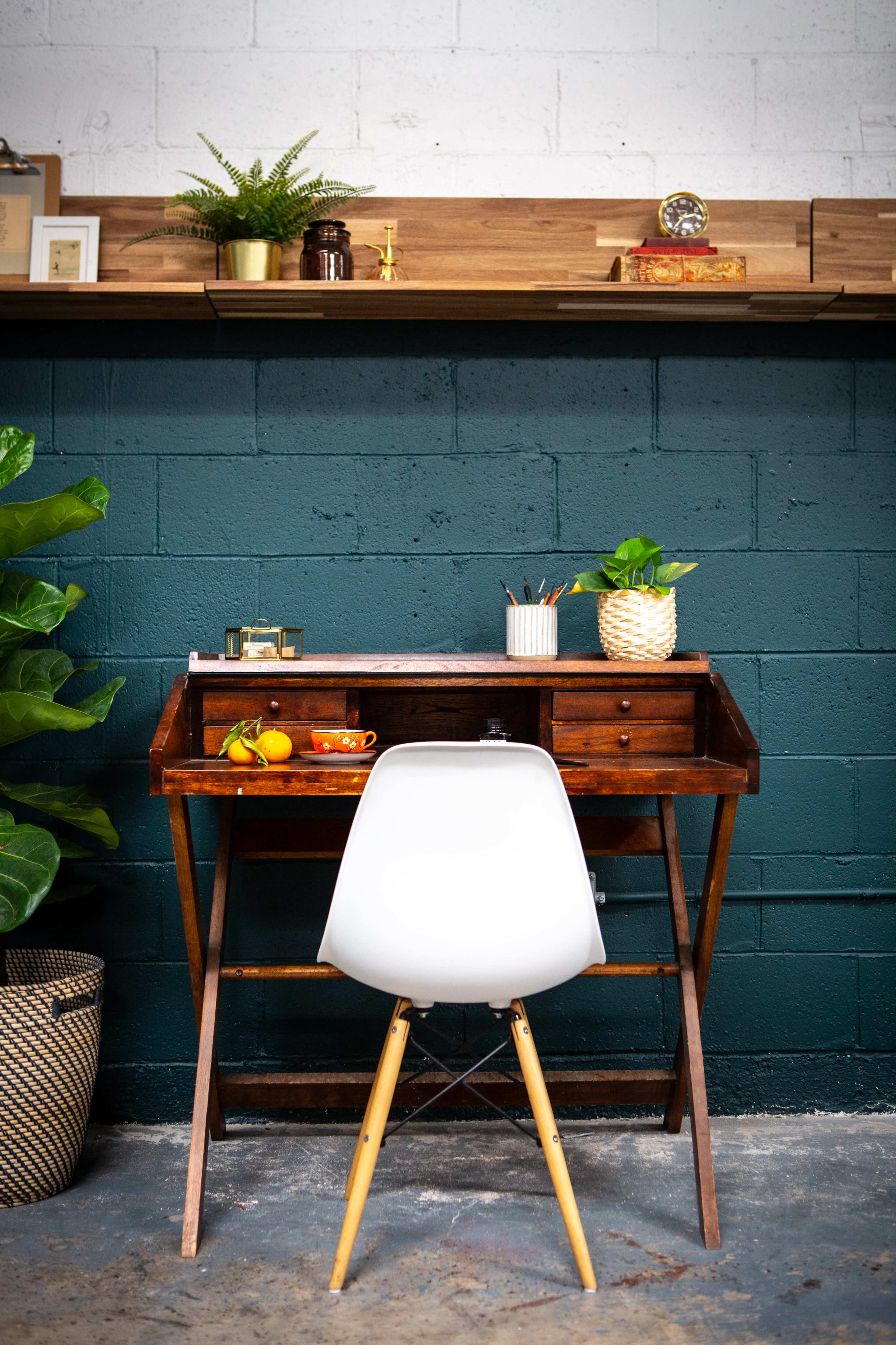 A wooden desk with a white chair is positioned against a dark green wall, adorned with potted plants and decorative items on a shelf above.