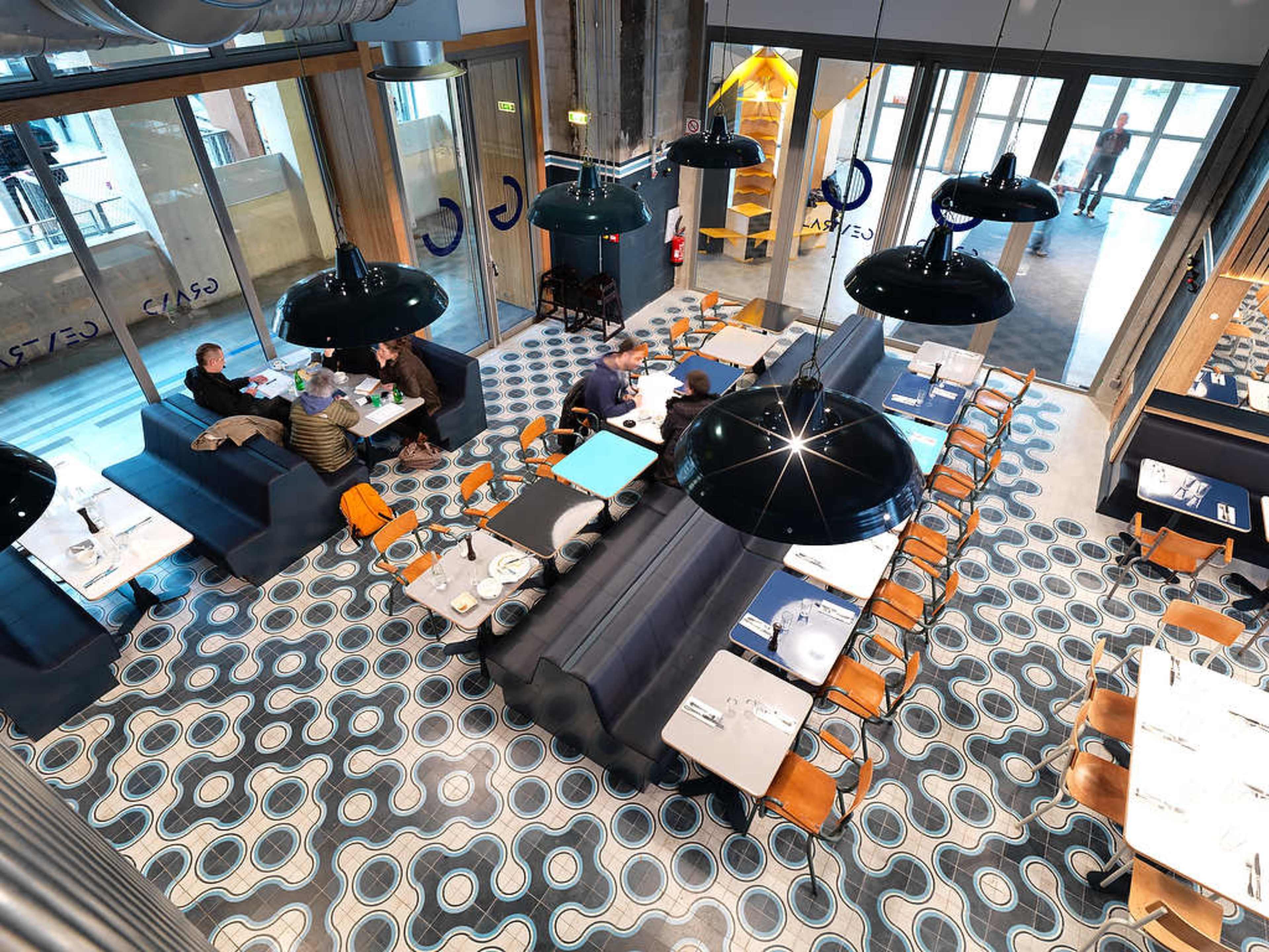 A modern restaurant interior with a patterned tiled floor, large black pendant lights, and a mix of tables and booths filled with diners.