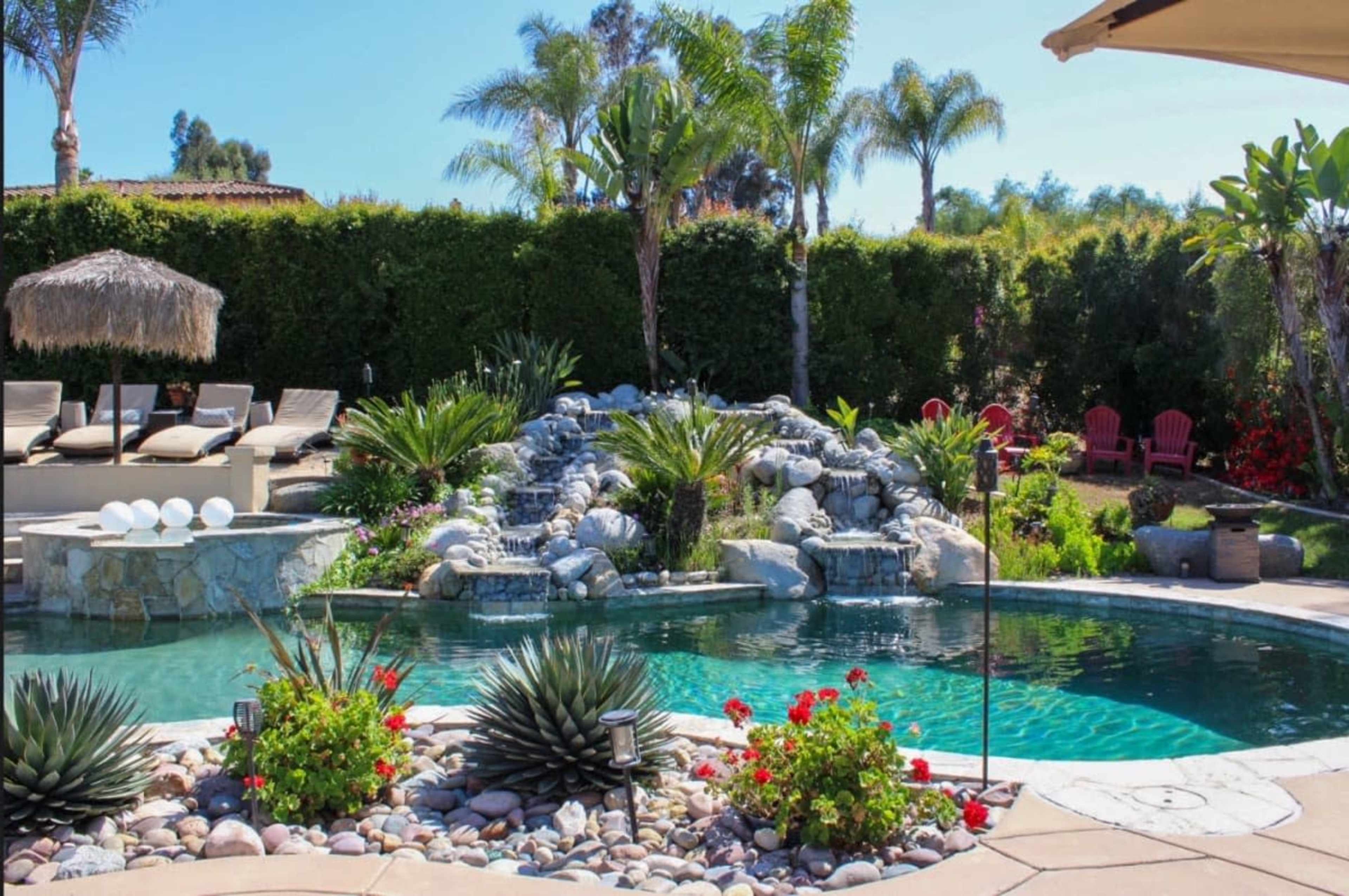 The image shows a swimming pool surrounded by tropical plants, a waterfall feature, and lounge chairs under a thatched umbrella.