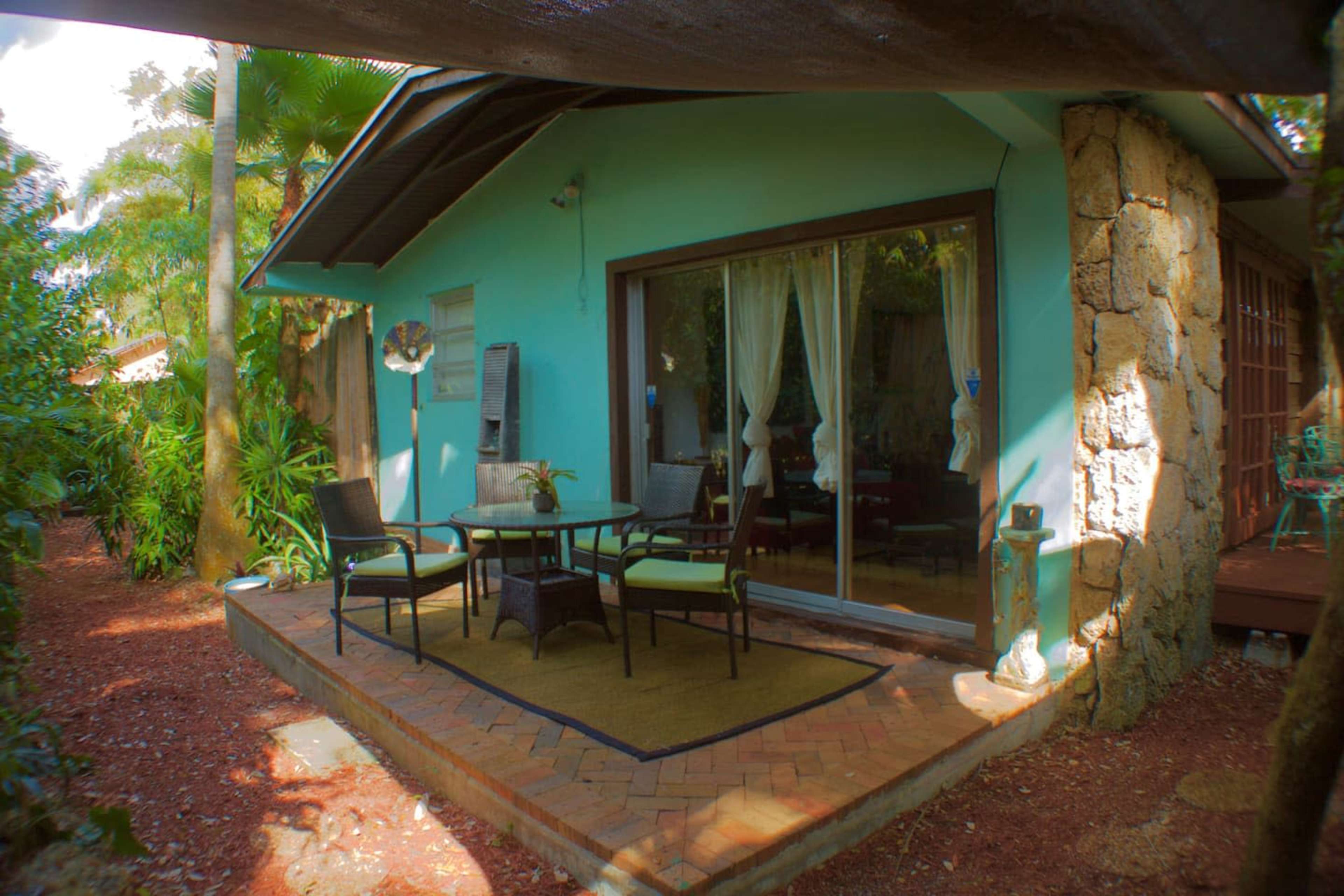 A small patio area with a table and chairs is situated outside a turquoise-colored building surrounded by greenery.