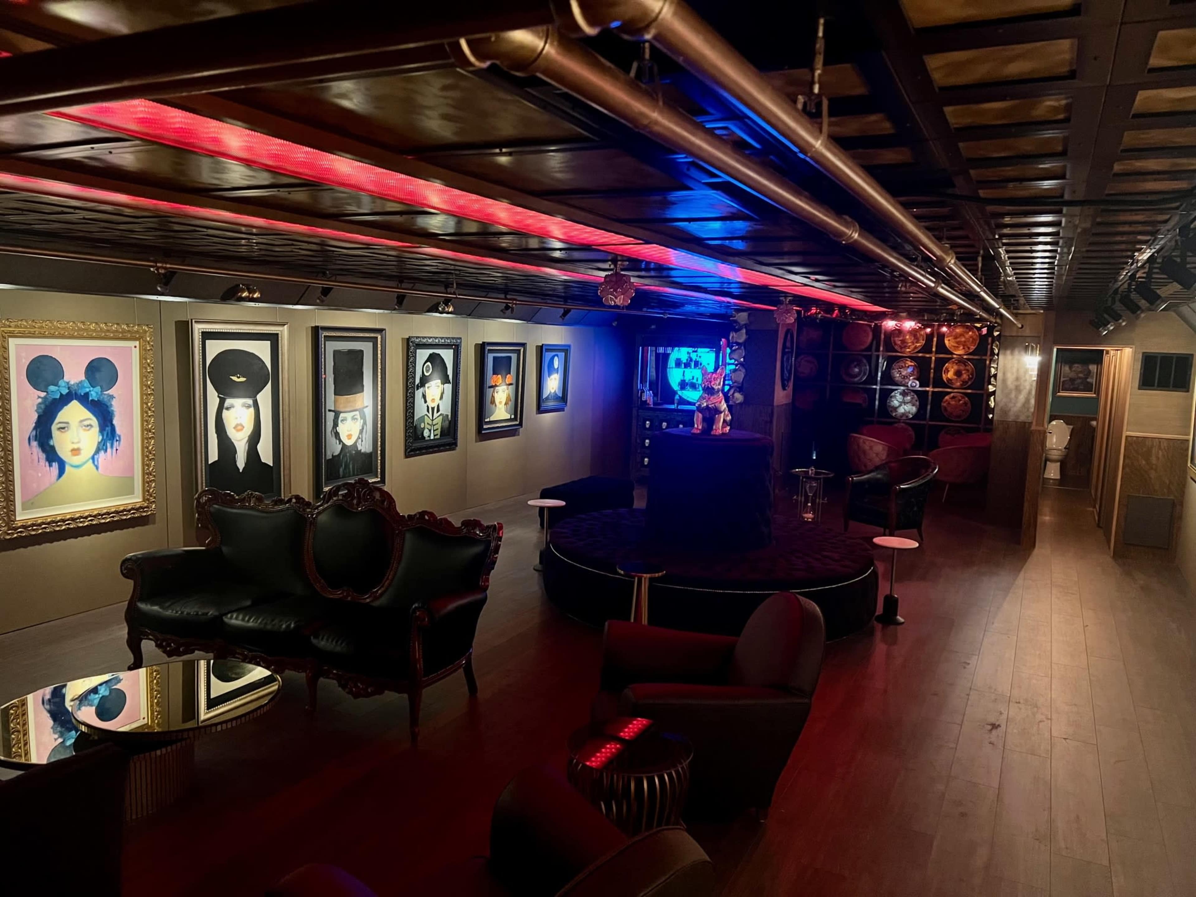 The image shows a dimly lit lounge with artwork on the walls and a central bar area surrounded by plush seating.