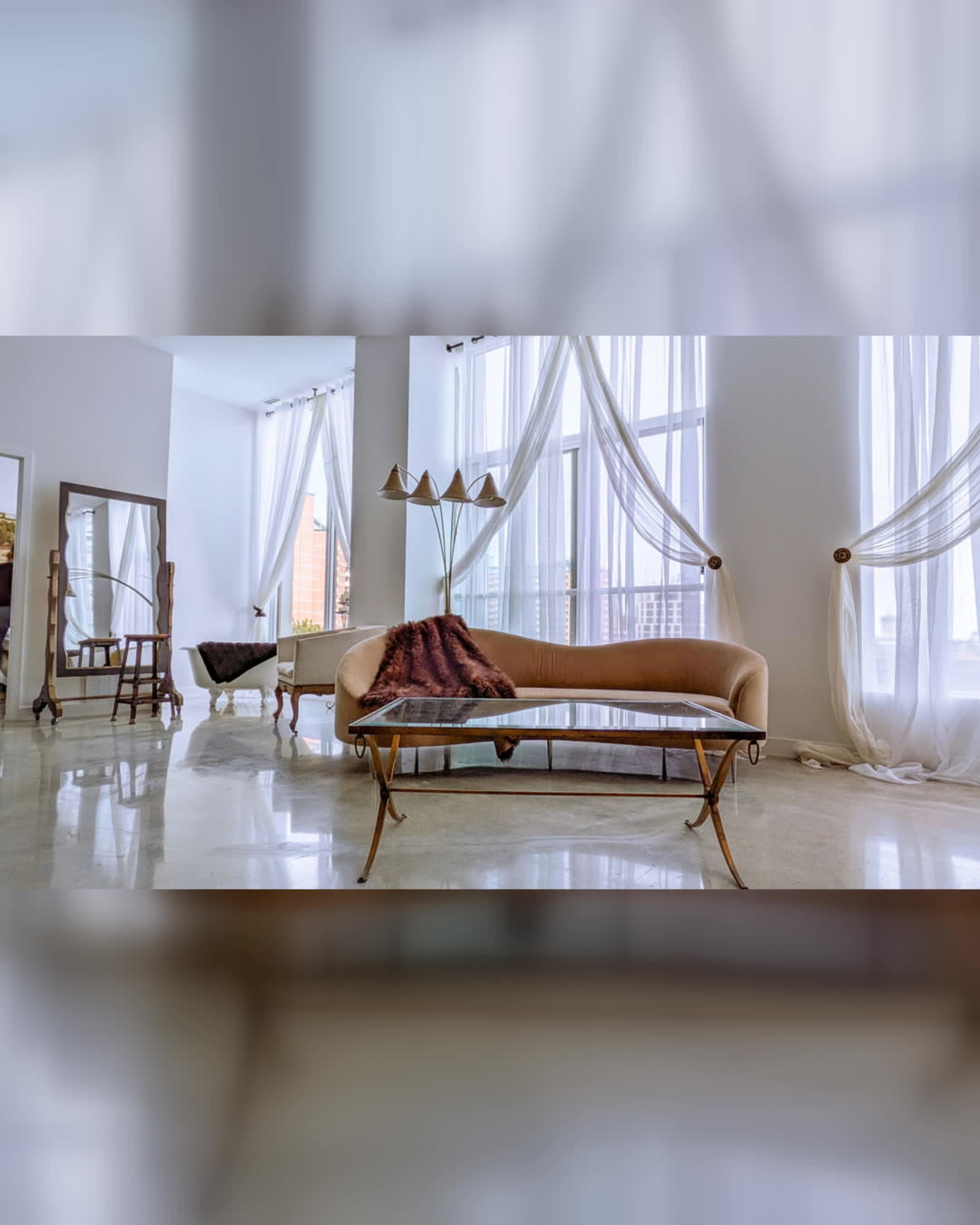 The image shows a modern living room featuring a curved sofa, a glass coffee table, and large windows draped with light fabric.