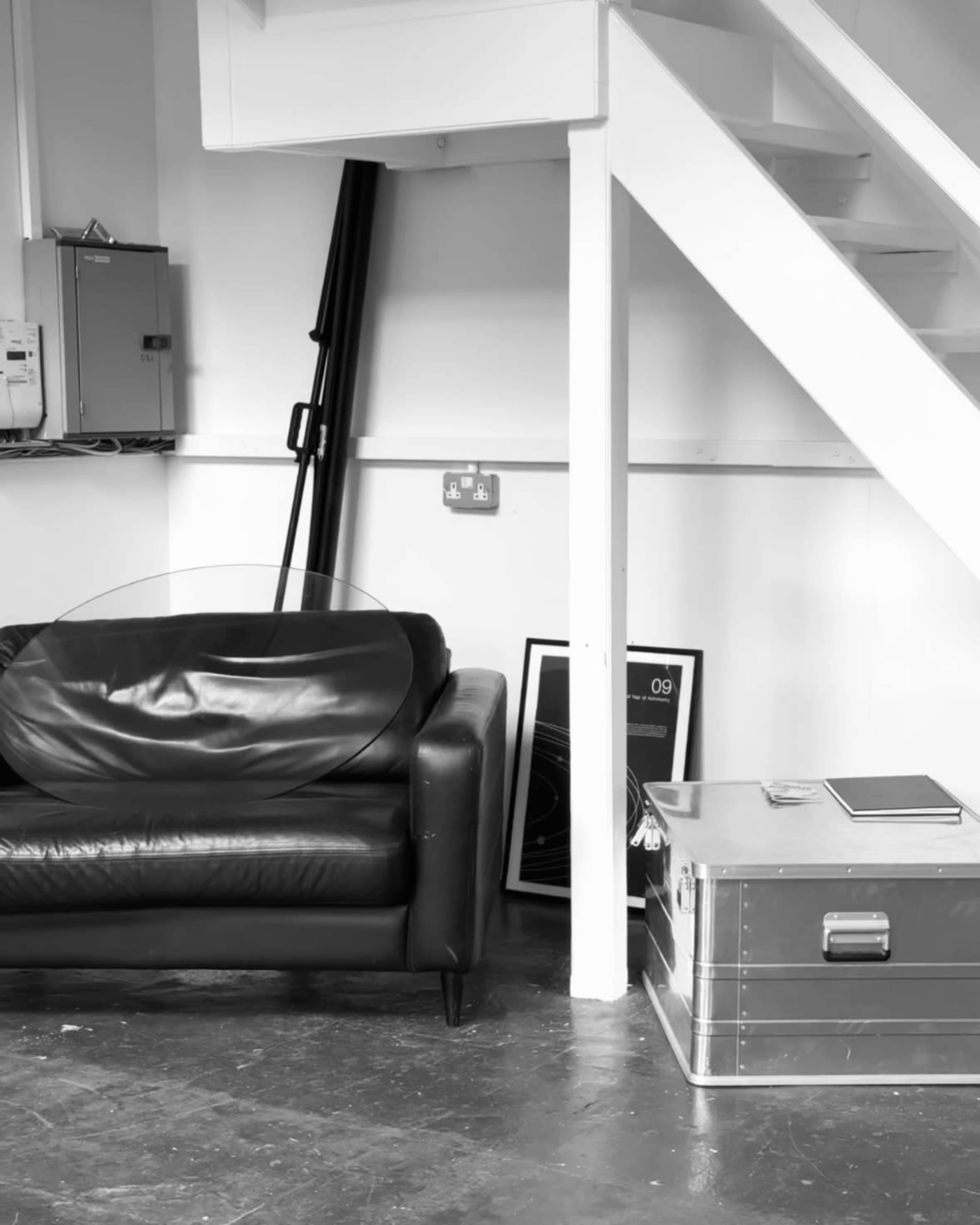 The image shows a black leather couch positioned near a white staircase, with a glass tabletop, a metallic storage box, and framed artwork nearby.