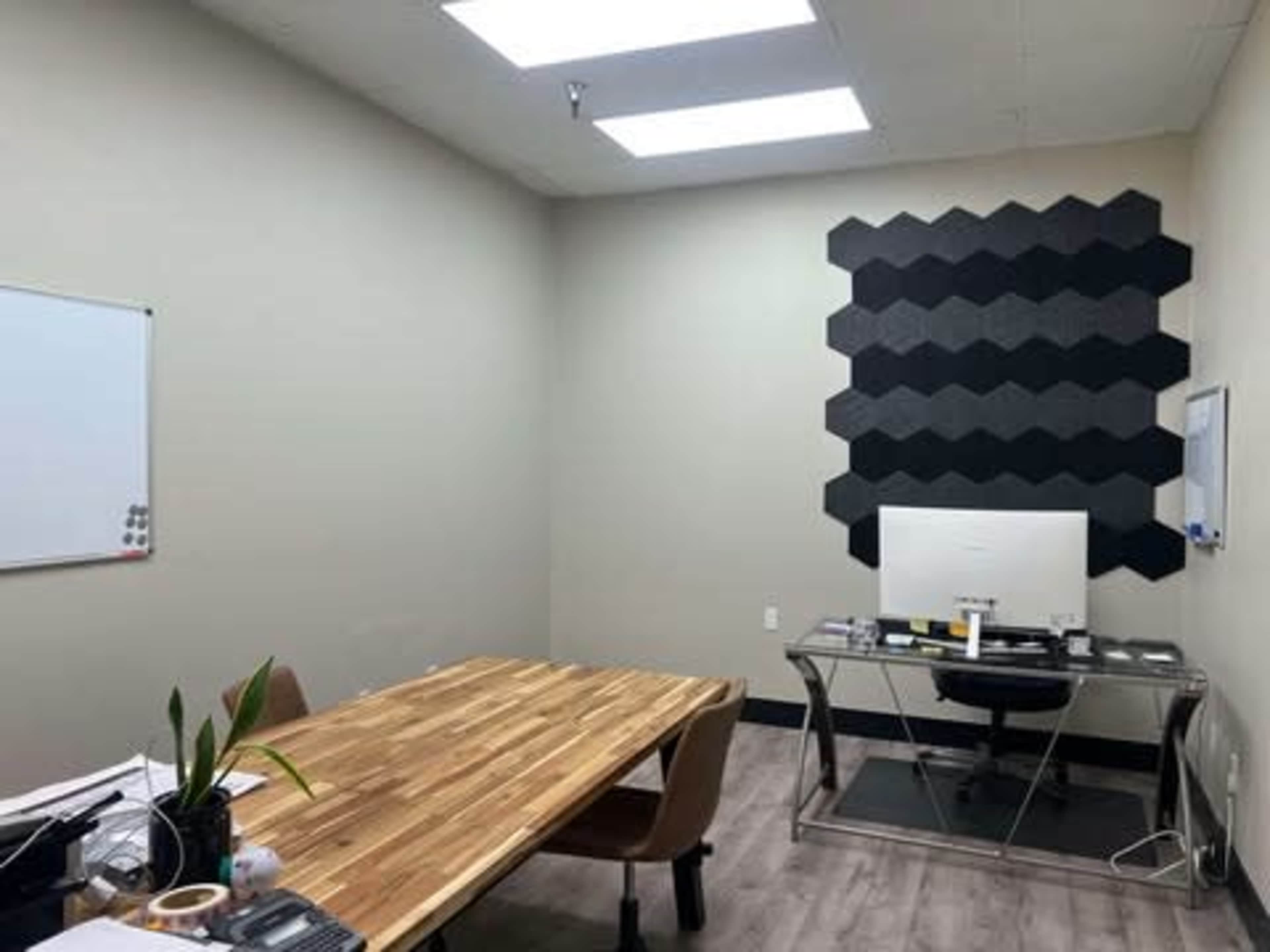 The image shows a minimalist office with a wooden table, a desk with a computer, and hexagonal sound panels on one wall.