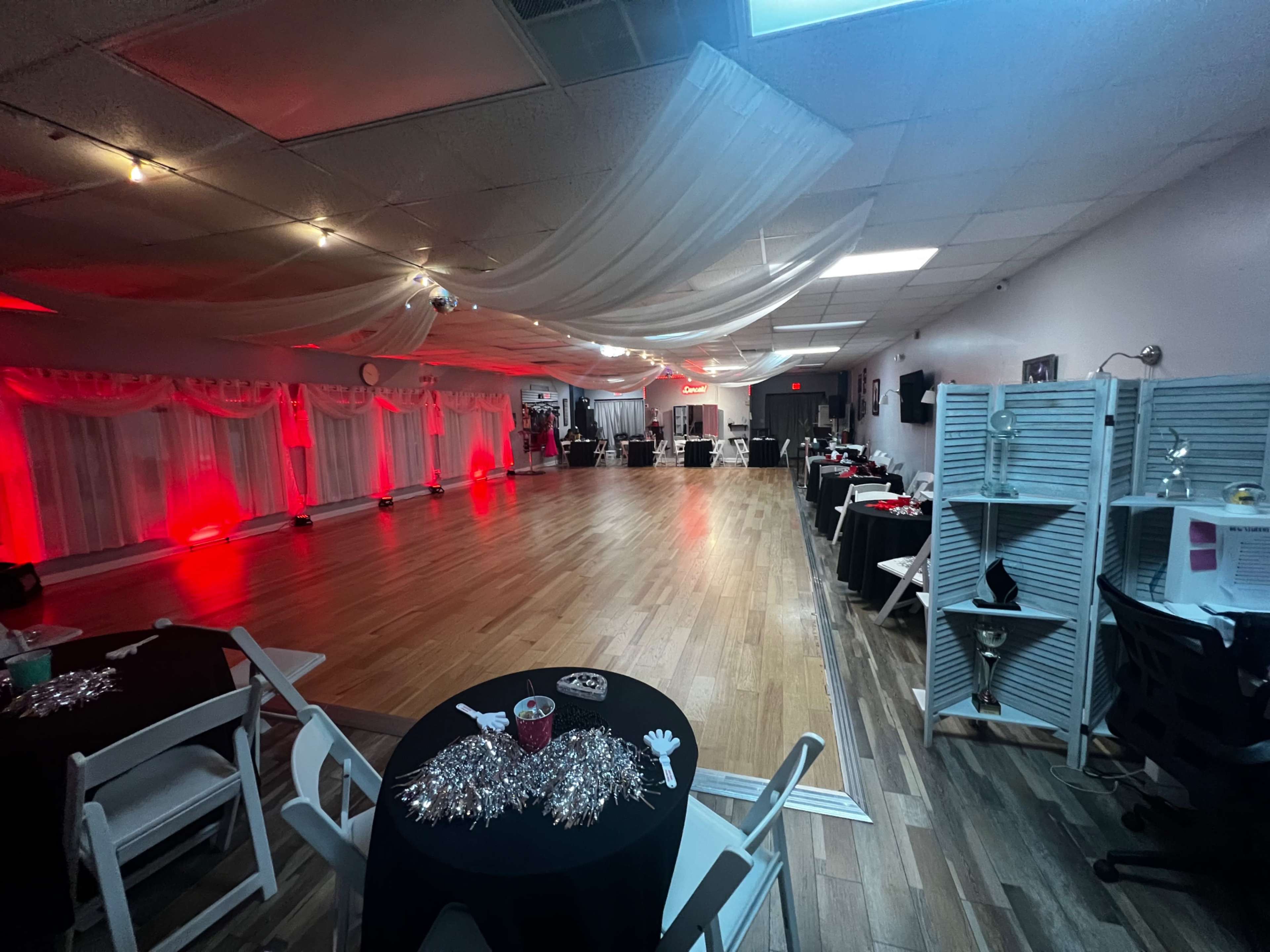 A spacious event hall with hardwood floors, stage lighting in red, and tables set up around the perimeter.