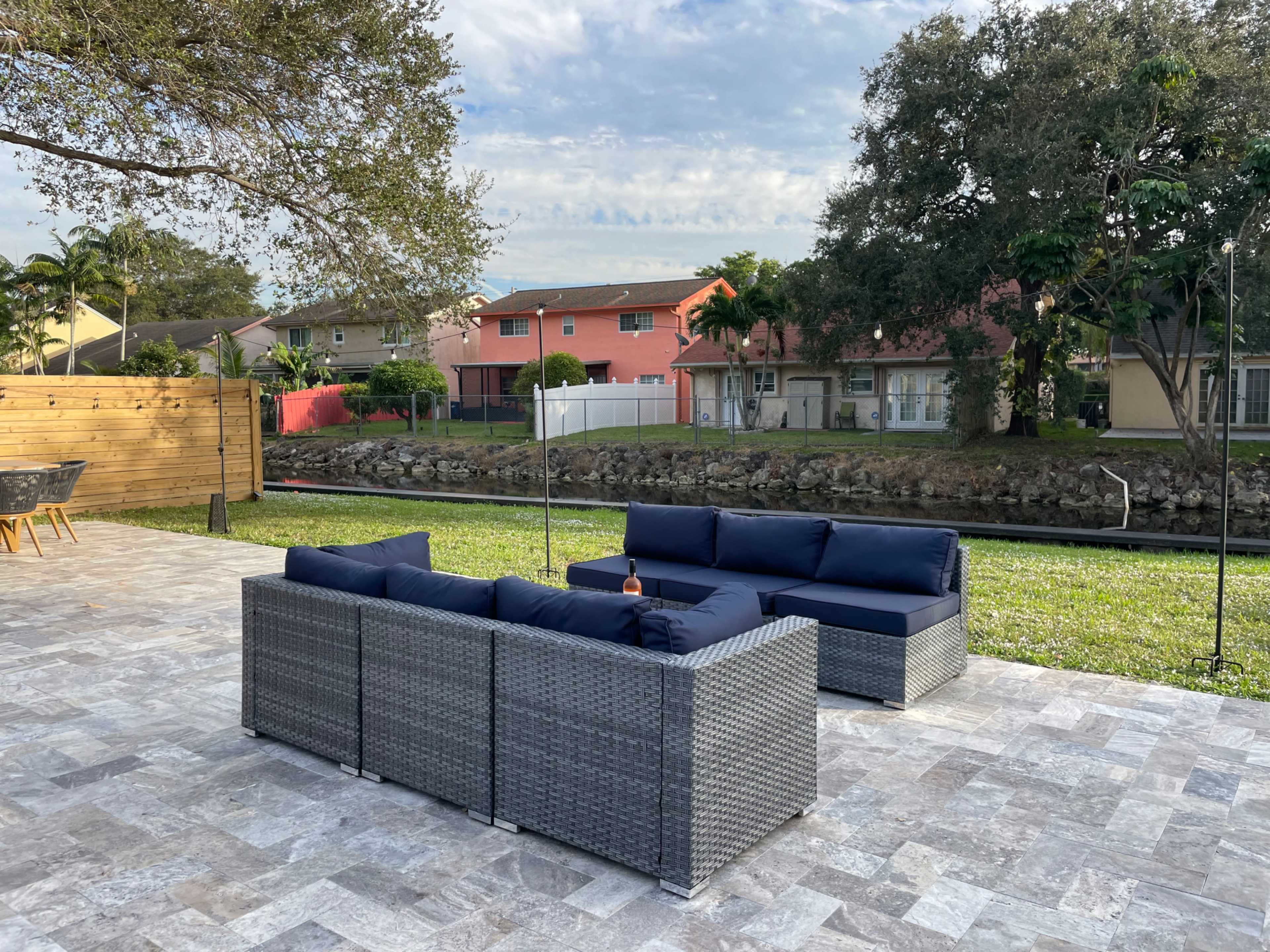 A modern outdoor seating area features a gray wicker couch with navy cushions, set against a backdrop of houses and greenery.