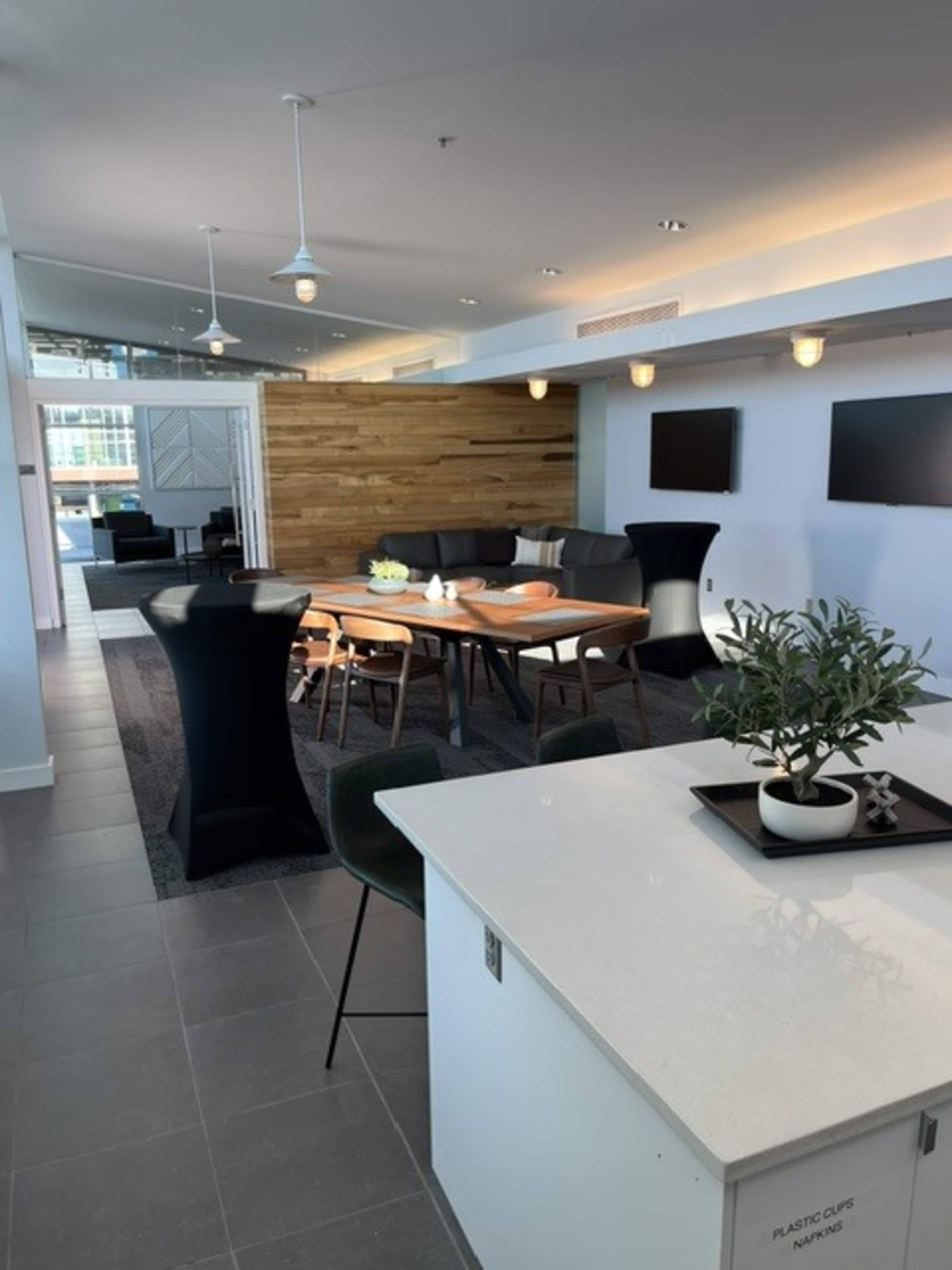 The image shows a modern interior space featuring a dining area with a wooden table, black chairs, and a living area with a sectional couch and two televisions mounted on the wall.