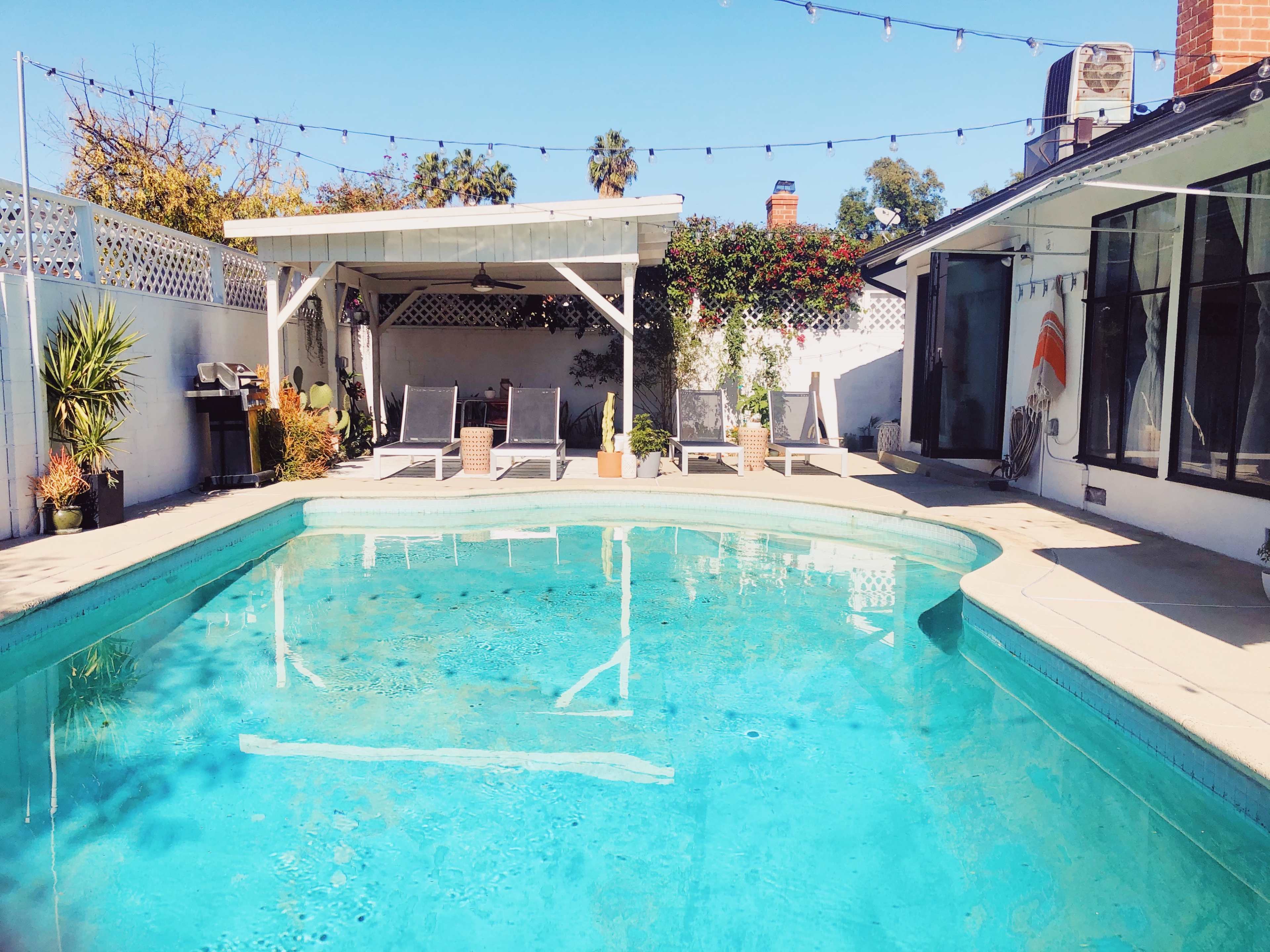 A swimming pool surrounded by lounge chairs, greenery, and a shaded patio area.