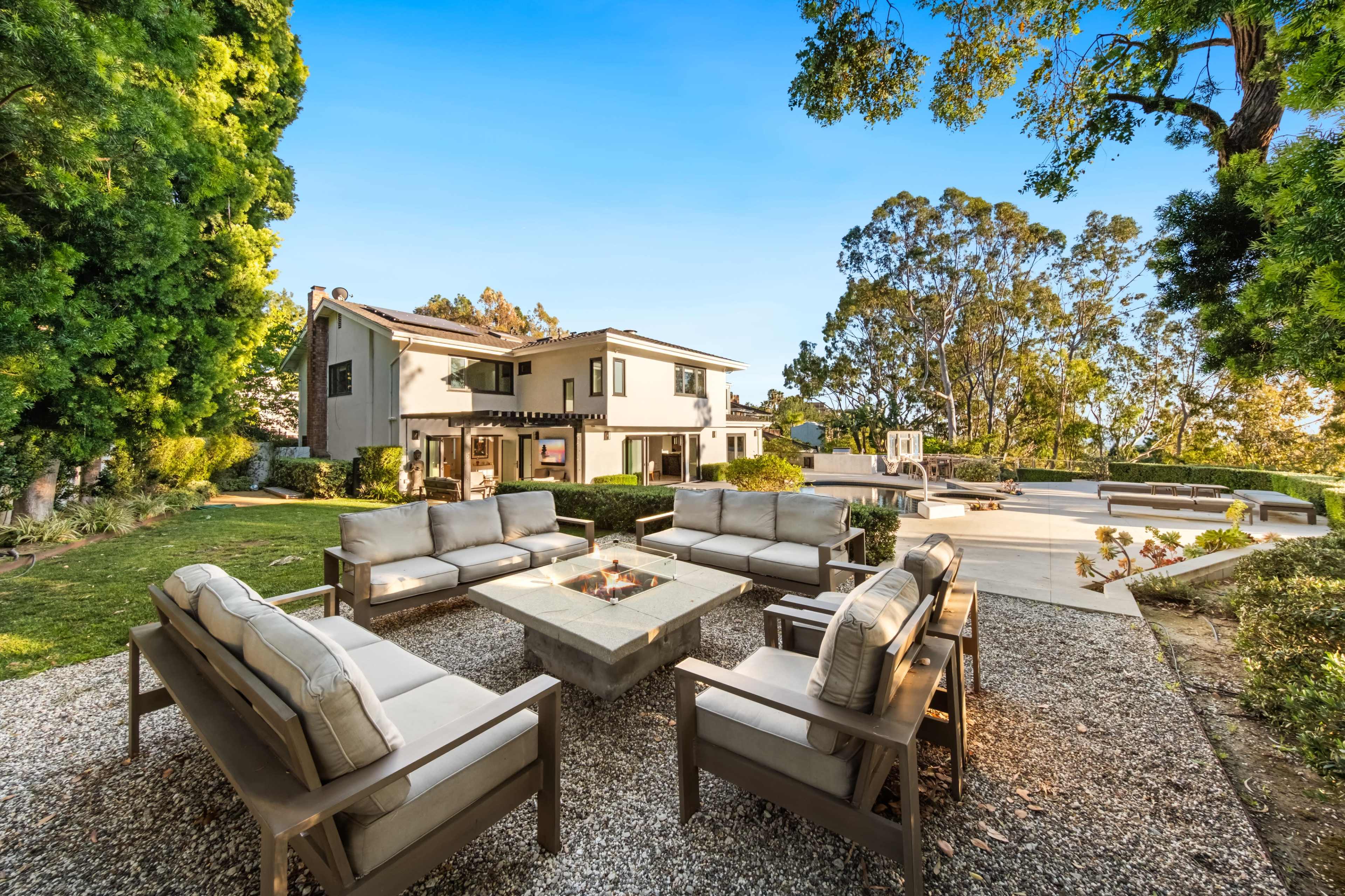 A modern backyard features a stone fire pit surrounded by several seating arrangements, with a spacious home and trees in the background.