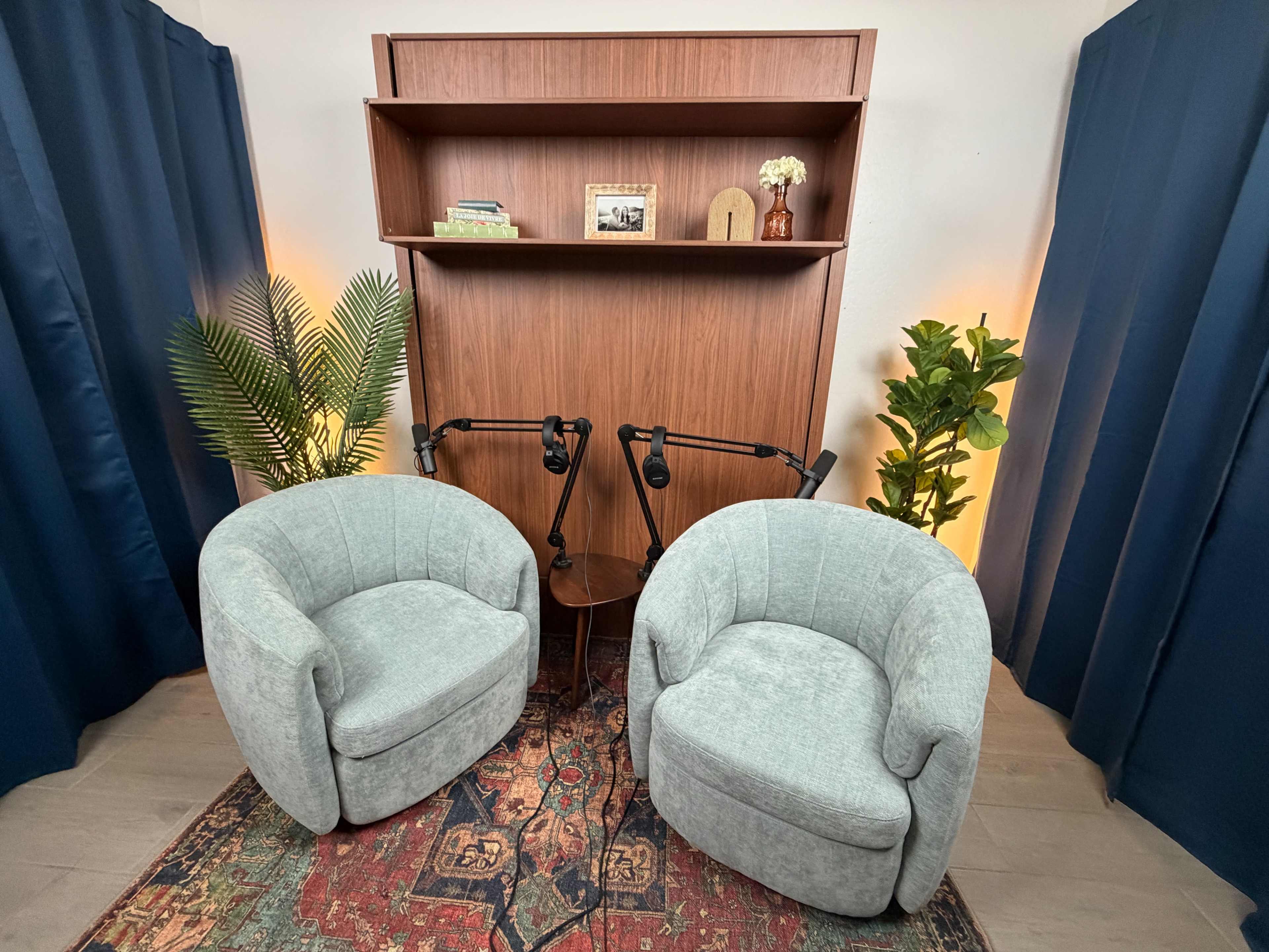 The image shows two gray, curved armchairs facing each other in front of a wooden bookshelf, with indoor plants on either side and two microphones attached to a small table between the chairs.