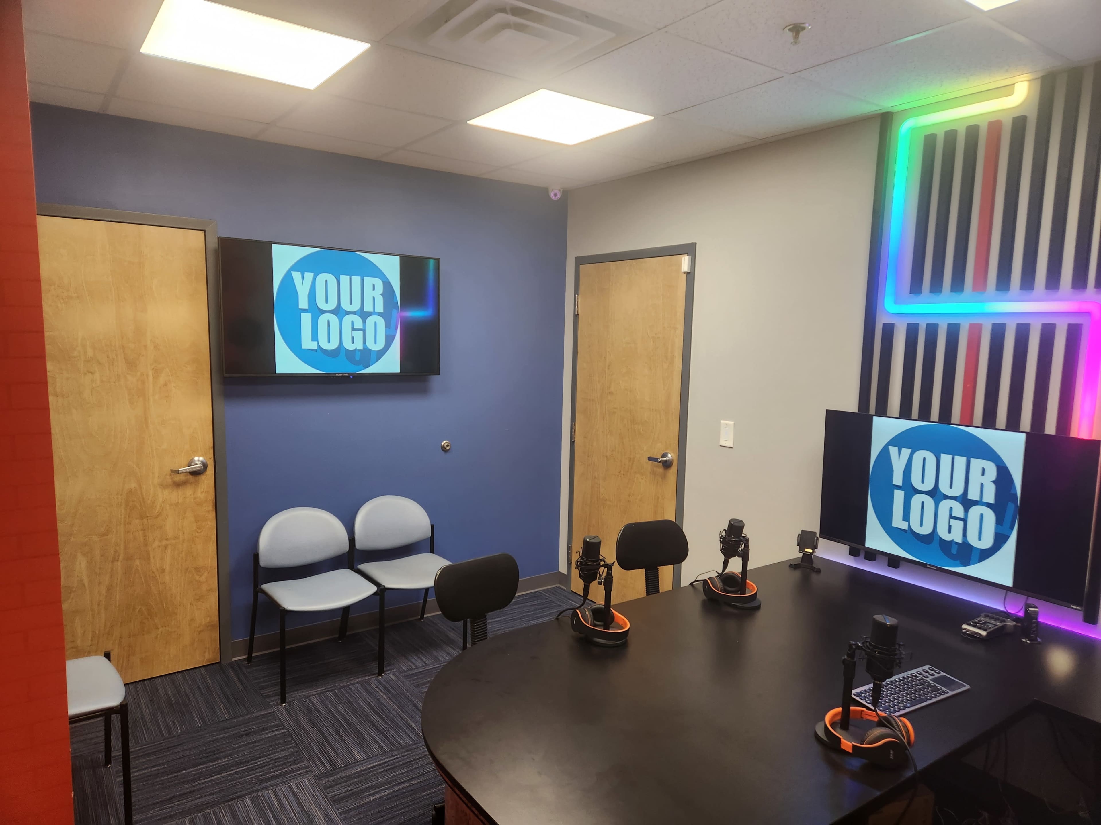 The image shows a modern recording studio with two televisions displaying "YOUR LOGO," a round table with microphones, and several chairs arranged nearby.