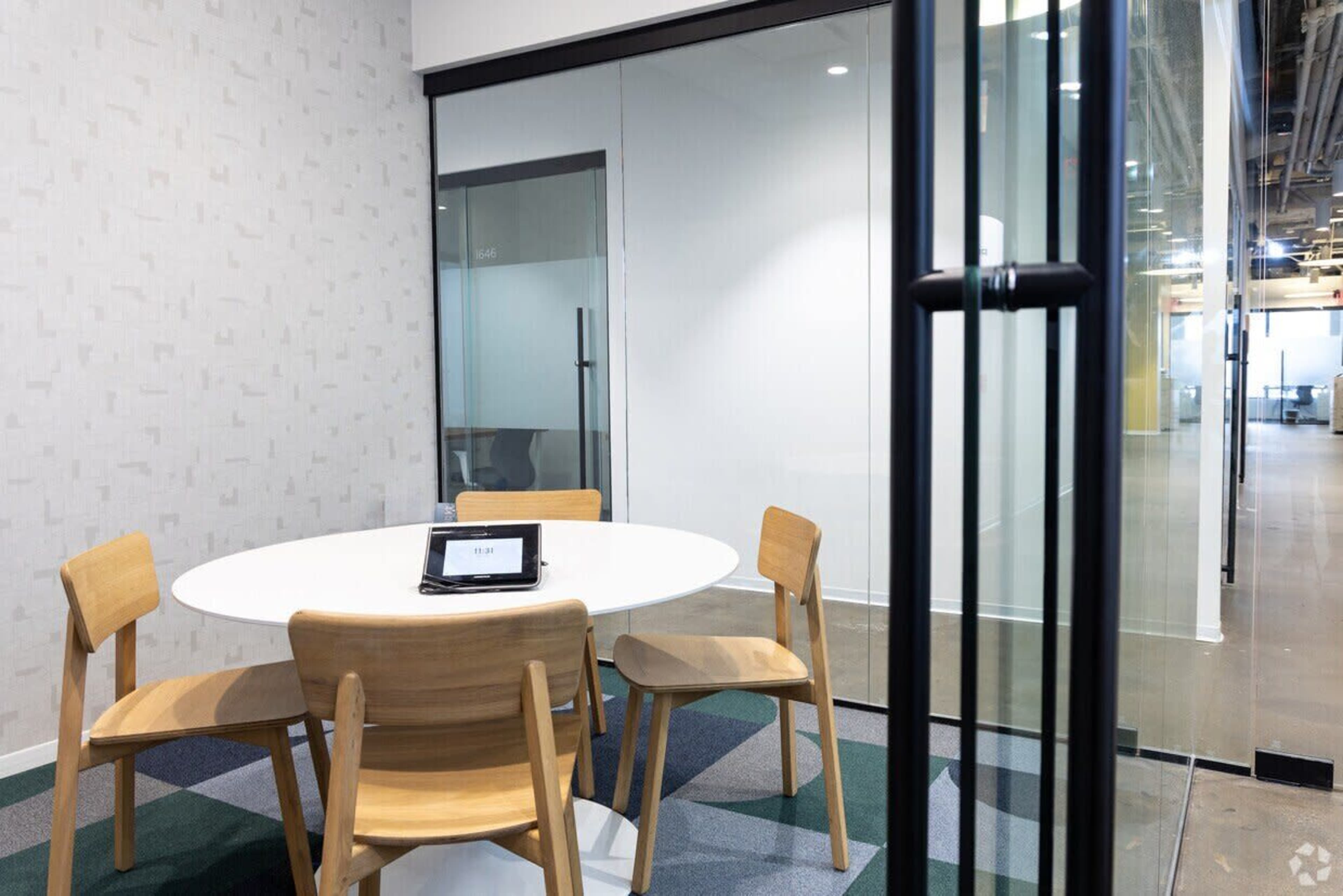 A small conference room features a round table with four wooden chairs, a digital screen on the table, and glass walls.