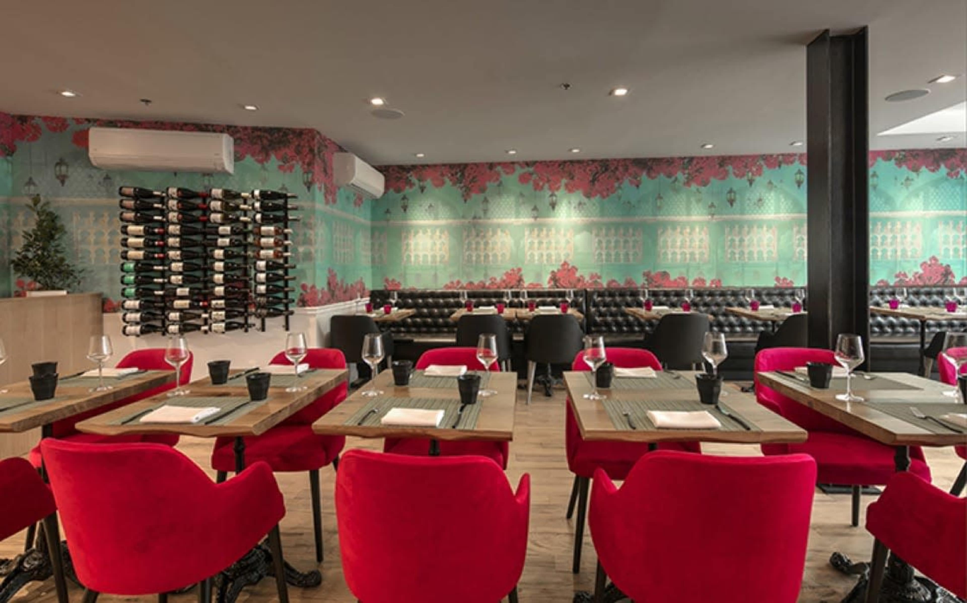 The image shows a stylish restaurant interior with red chairs, wooden tables, and a decorative wall adorned with floral wallpaper and a wine display.