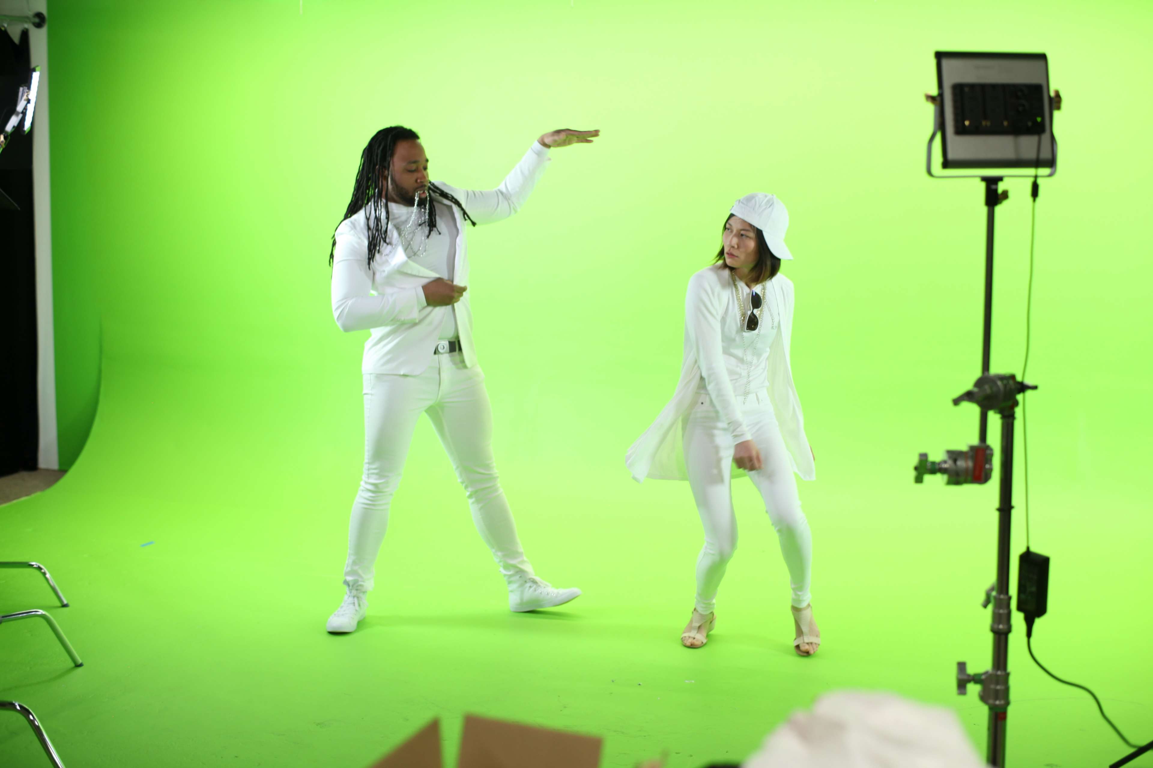 Two individuals in white attire perform in front of a bright green backdrop.