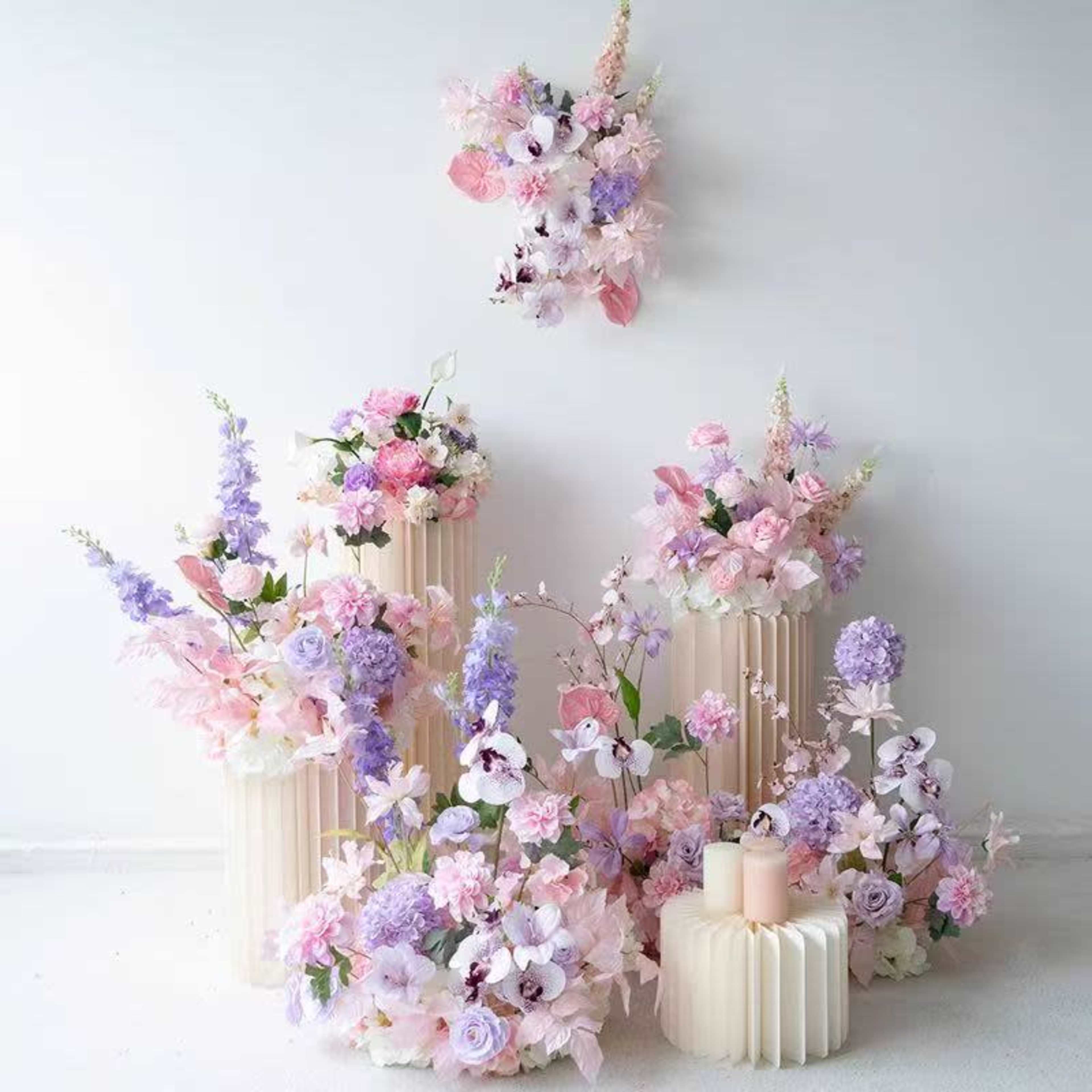 The image features an artistic floral arrangement with pastel-colored flowers displayed on several cream-colored pedestals, complemented by a hanging floral piece above.