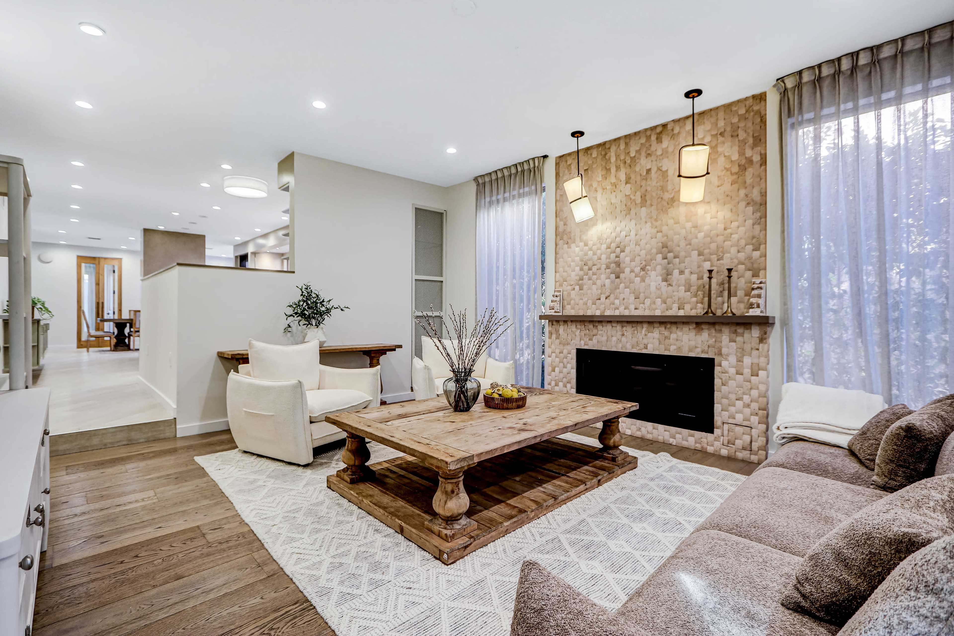 The image shows a modern living room featuring a comfortable seating area with a coffee table, a stone-patterned feature wall, and large windows covered with sheer curtains.