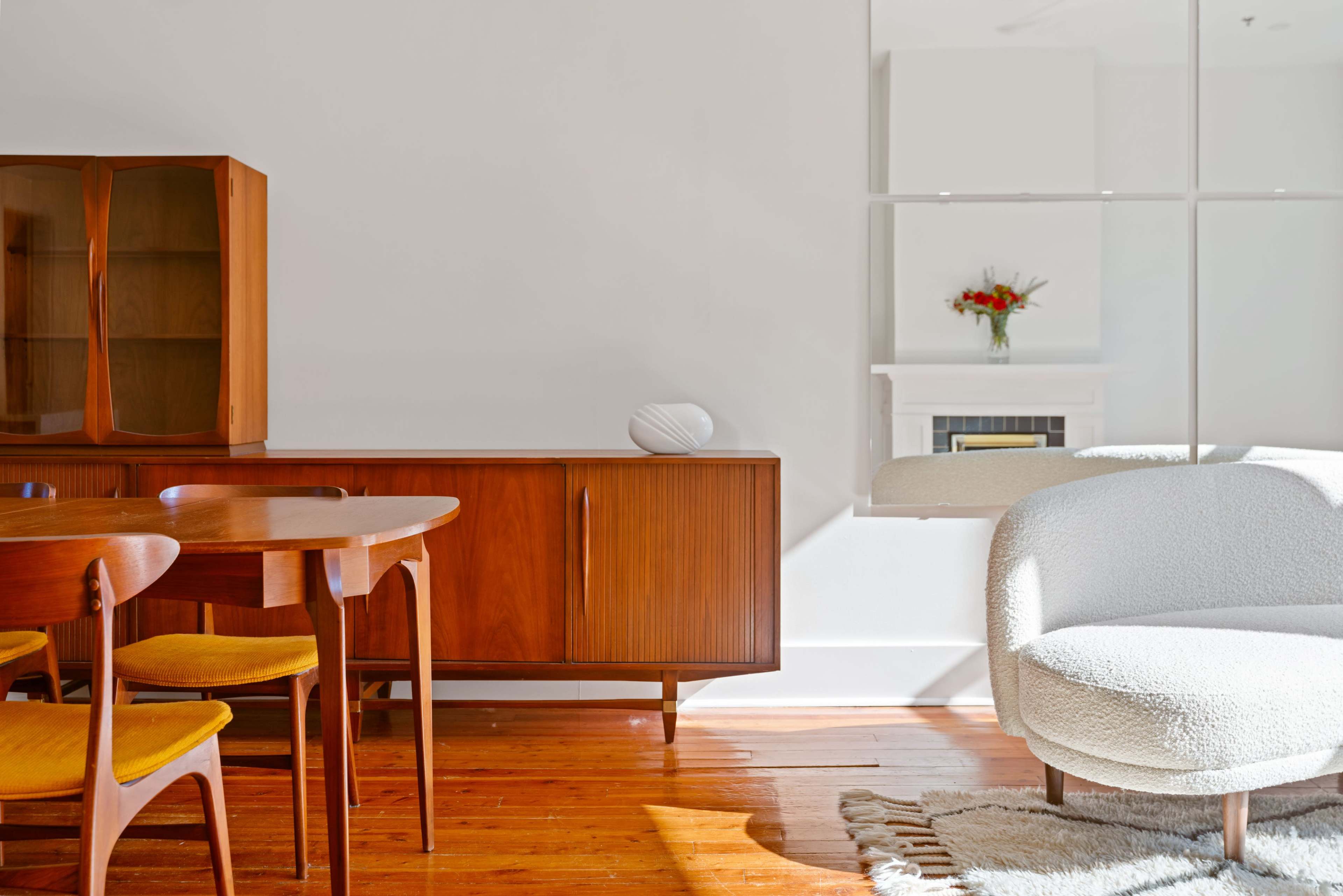 A mid-century modern dining area with a wooden table and chairs, a sideboard, and a cozy white armchair, all set against a light-colored wall.