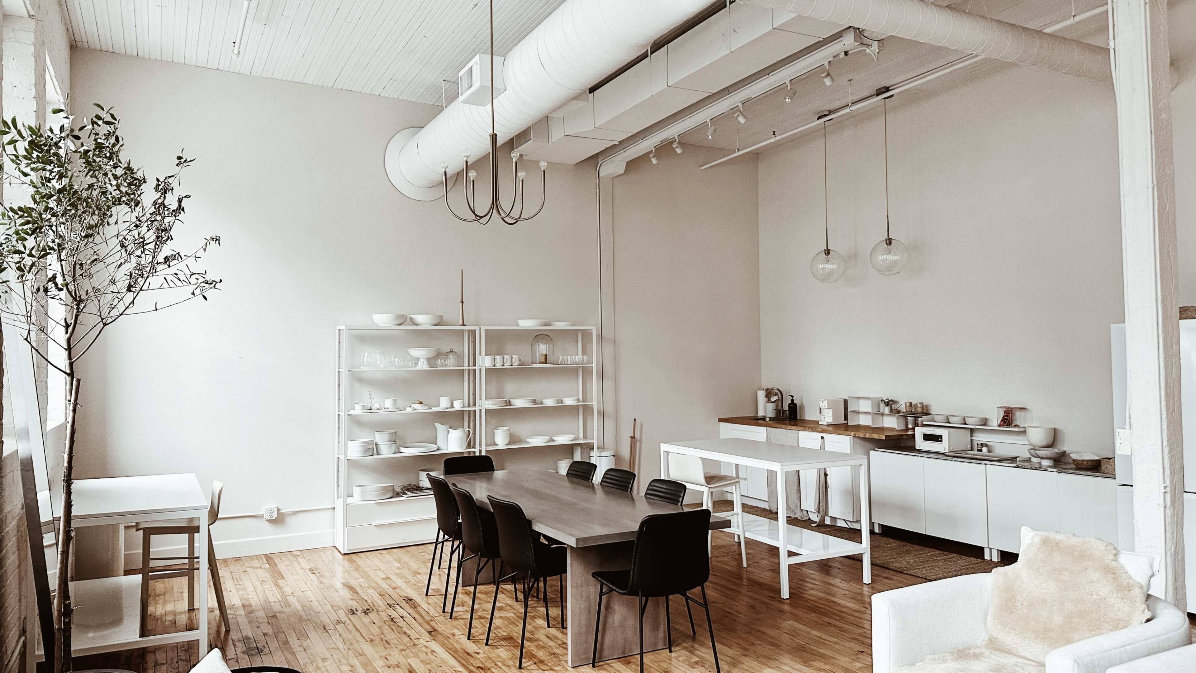 A minimalist kitchen and dining area with a long wooden table, black chairs, and shelves filled with white dishware.