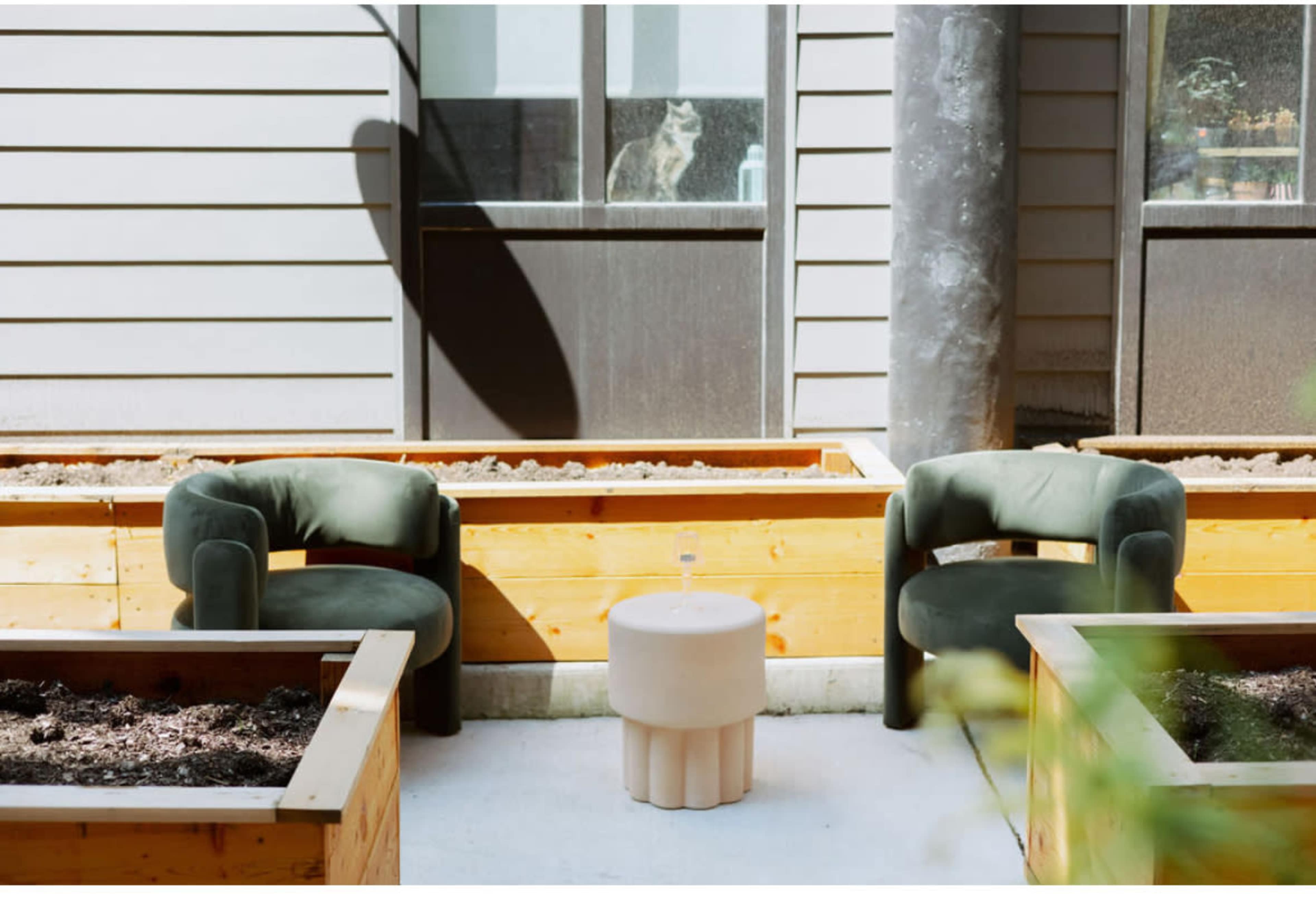 The image shows two green velvet chairs positioned near a circular stone table in a garden area with wooden planter boxes.
