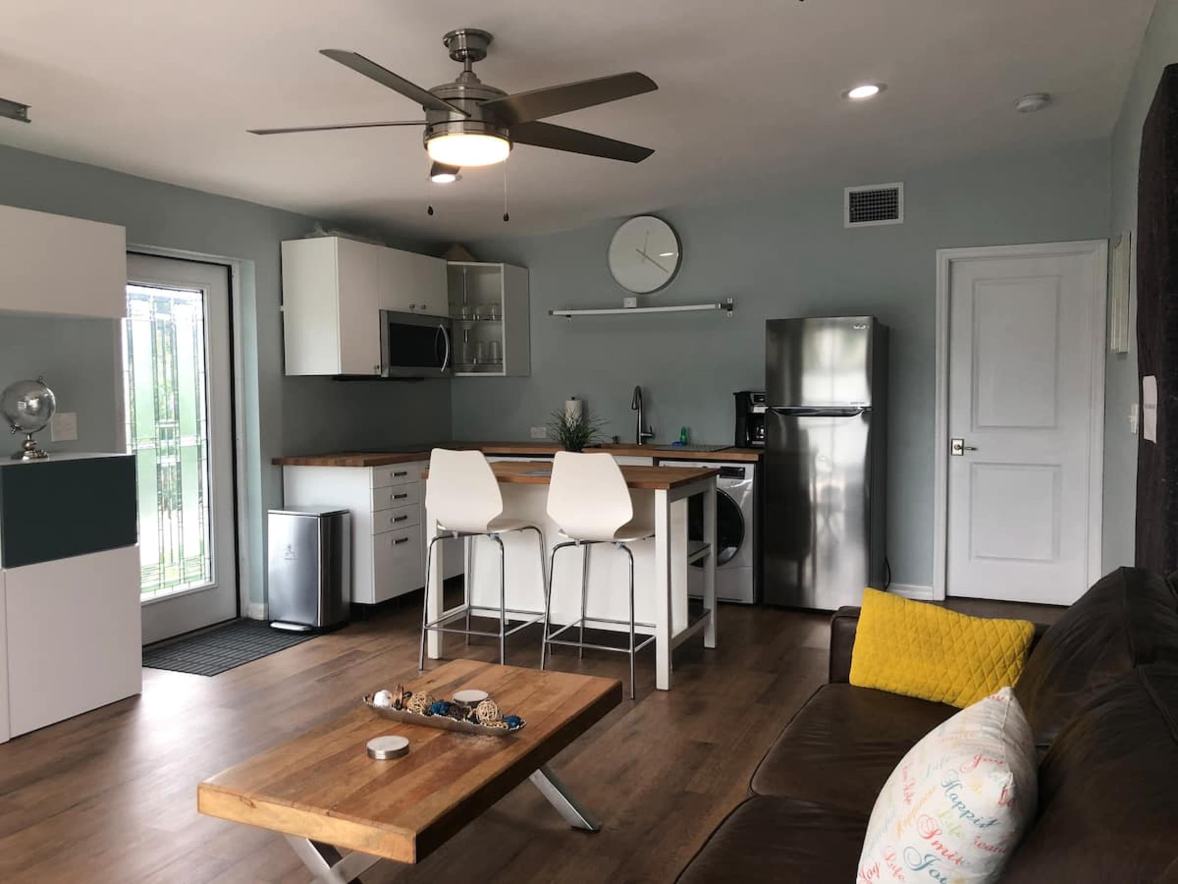 A modern kitchen and living area with a wooden table, white cabinetry, stainless steel appliances, and a ceiling fan.