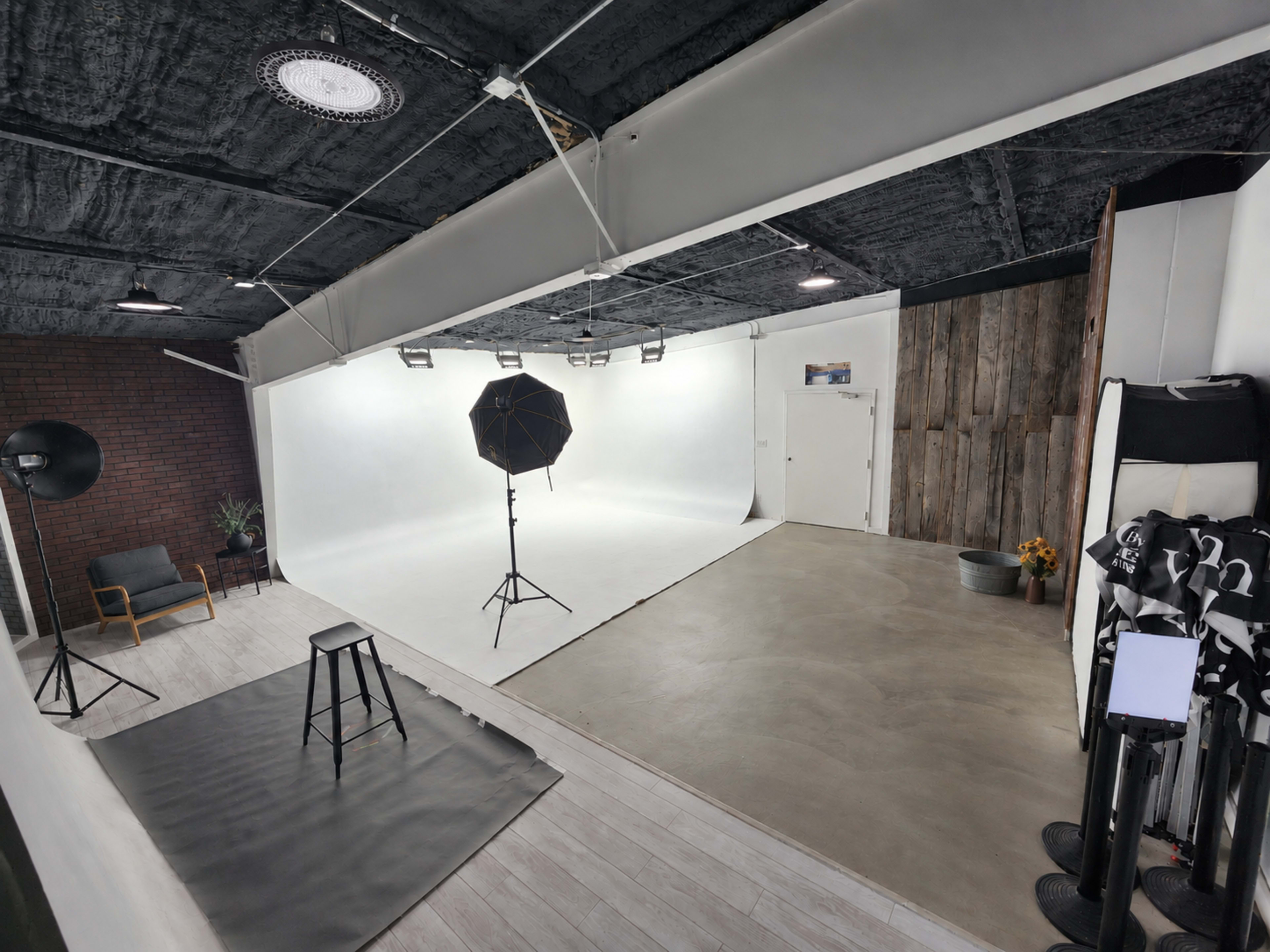 The image shows a photography studio featuring a white backdrop, lighting equipment, and a mix of concrete and wooden surfaces.