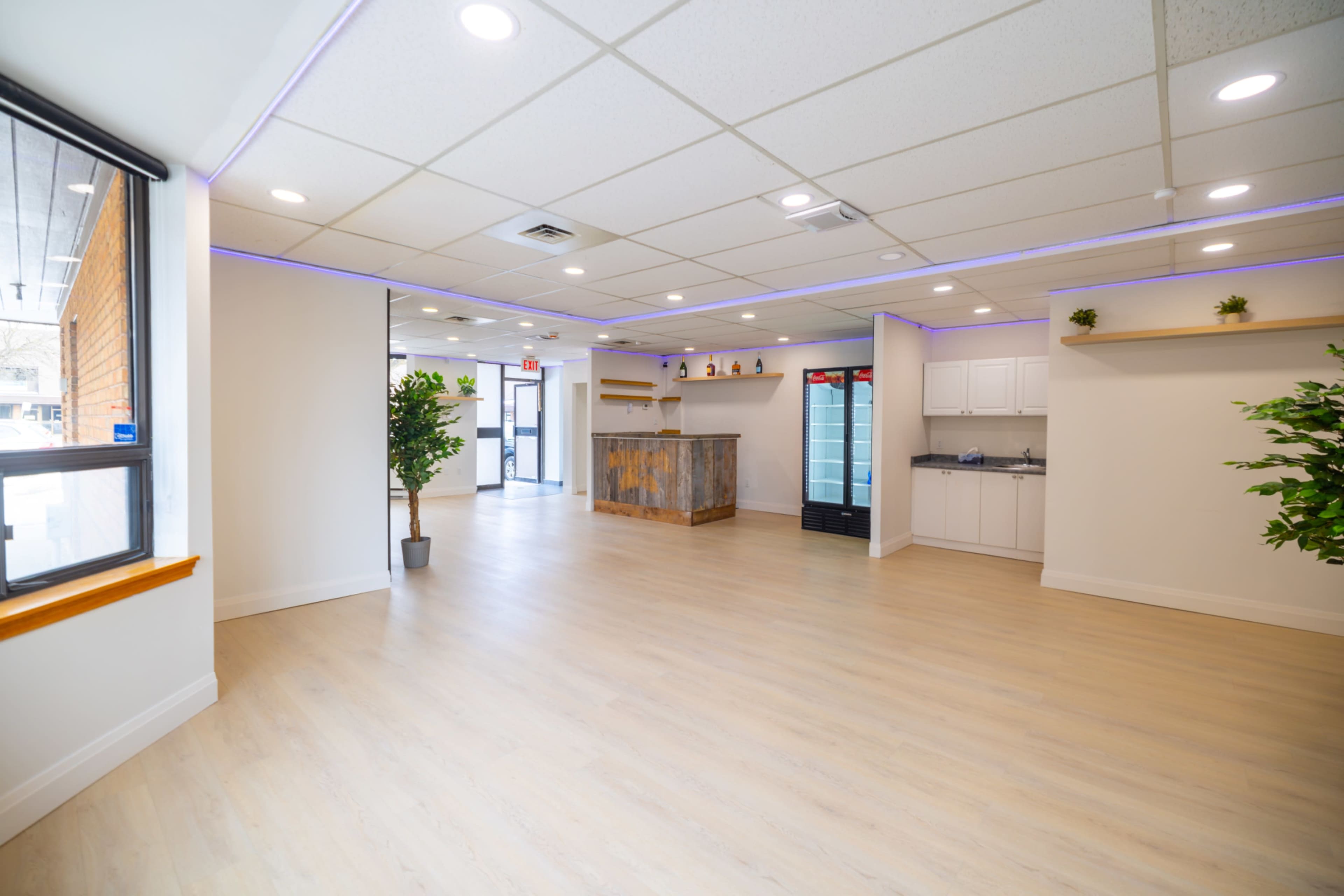 The image shows a spacious, well-lit interior with light wood flooring, a reception area featuring a wooden counter, and plants in the corners.
