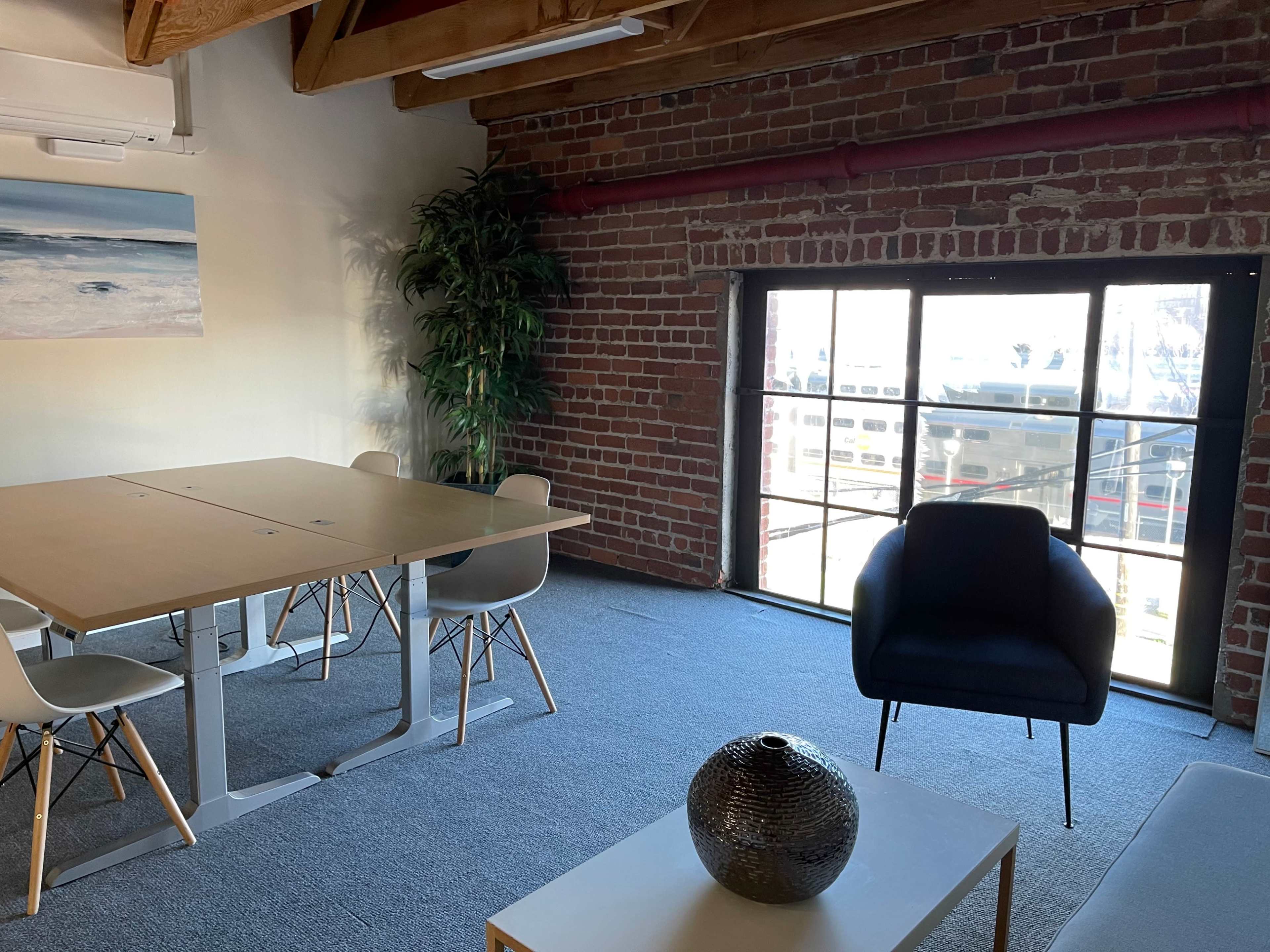 A modern office space with a wooden ceiling, exposed brick walls, a large window overlooking boats, a table with chairs, and a black armchair.