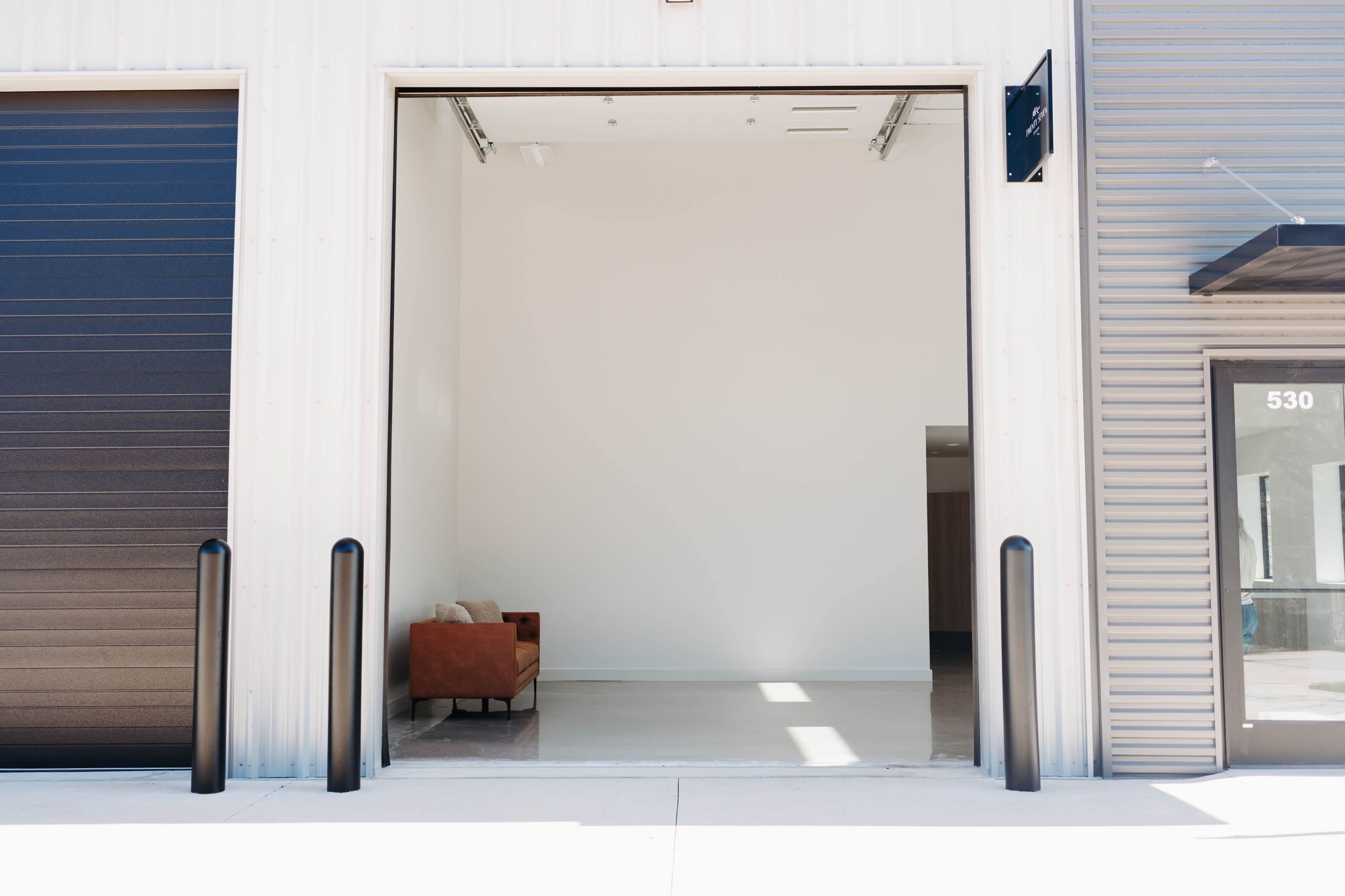 A large open garage door reveals a minimalist interior space featuring a single brown chair against a white wall.
