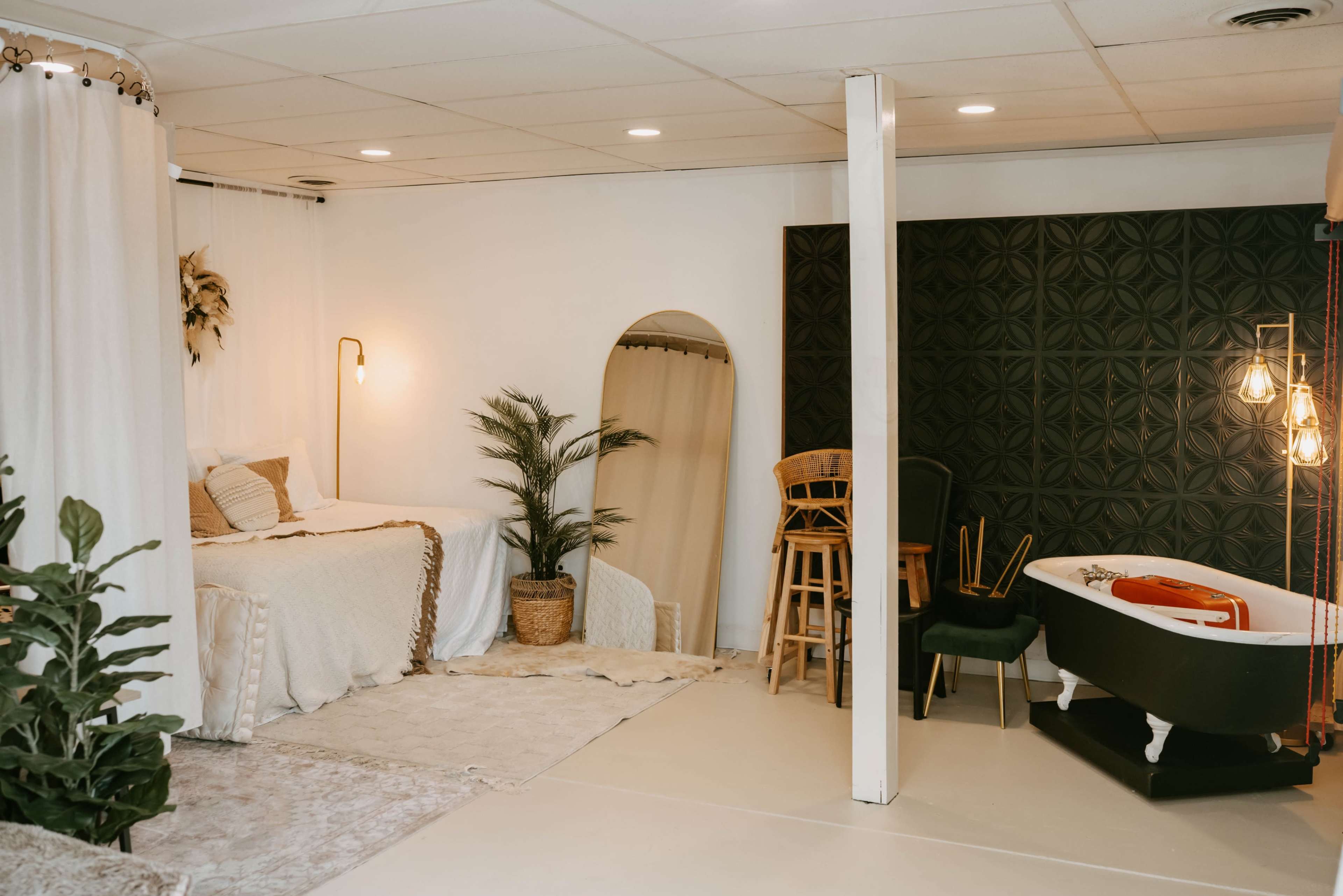 The image depicts a styled room featuring a bed with decorative pillows, a small plant in a woven basket, a bathtub with a red interior, and various seating arrangements against a textured wall.